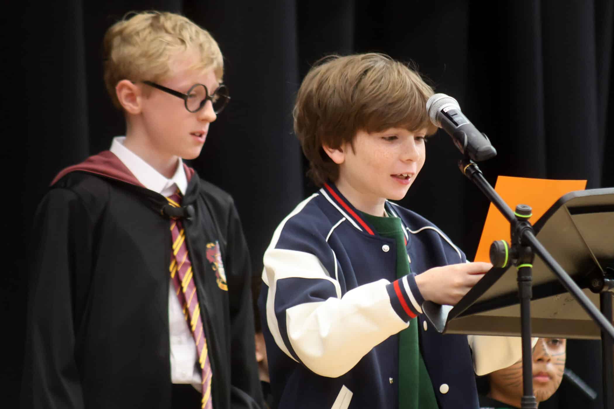 Two boys stand at a microphone. One wears glasses and a costume resembling a Harry Potter robe and tie; the other, in a jacket, reads from an orange paper. A black curtain is in the background.