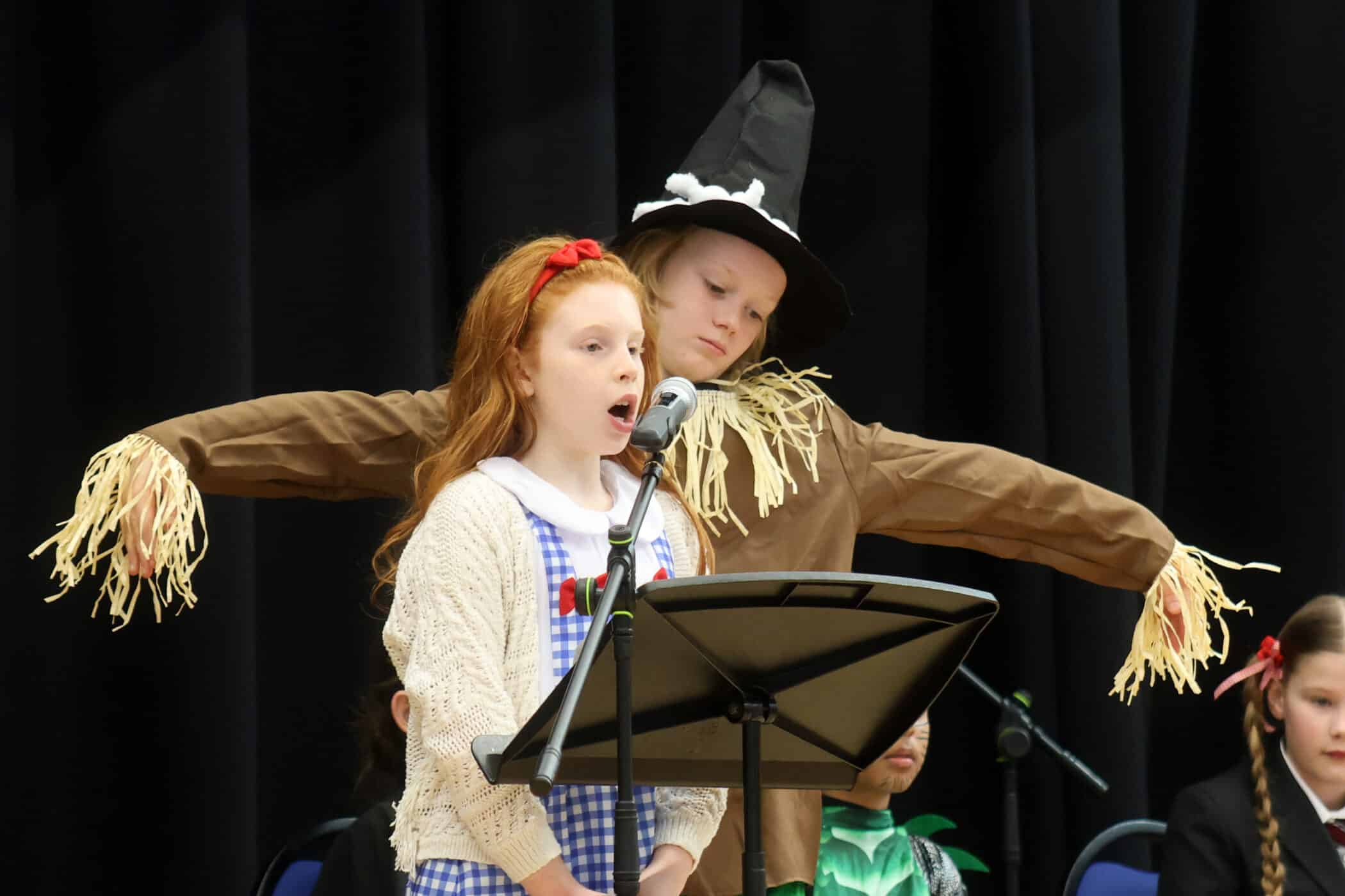 Two children perform on stage; one sings into a microphone wearing a gingham dress and cardigan, whilst the other, dressed as a scarecrow with a hat and straw details, stands behind with arms outstretched.