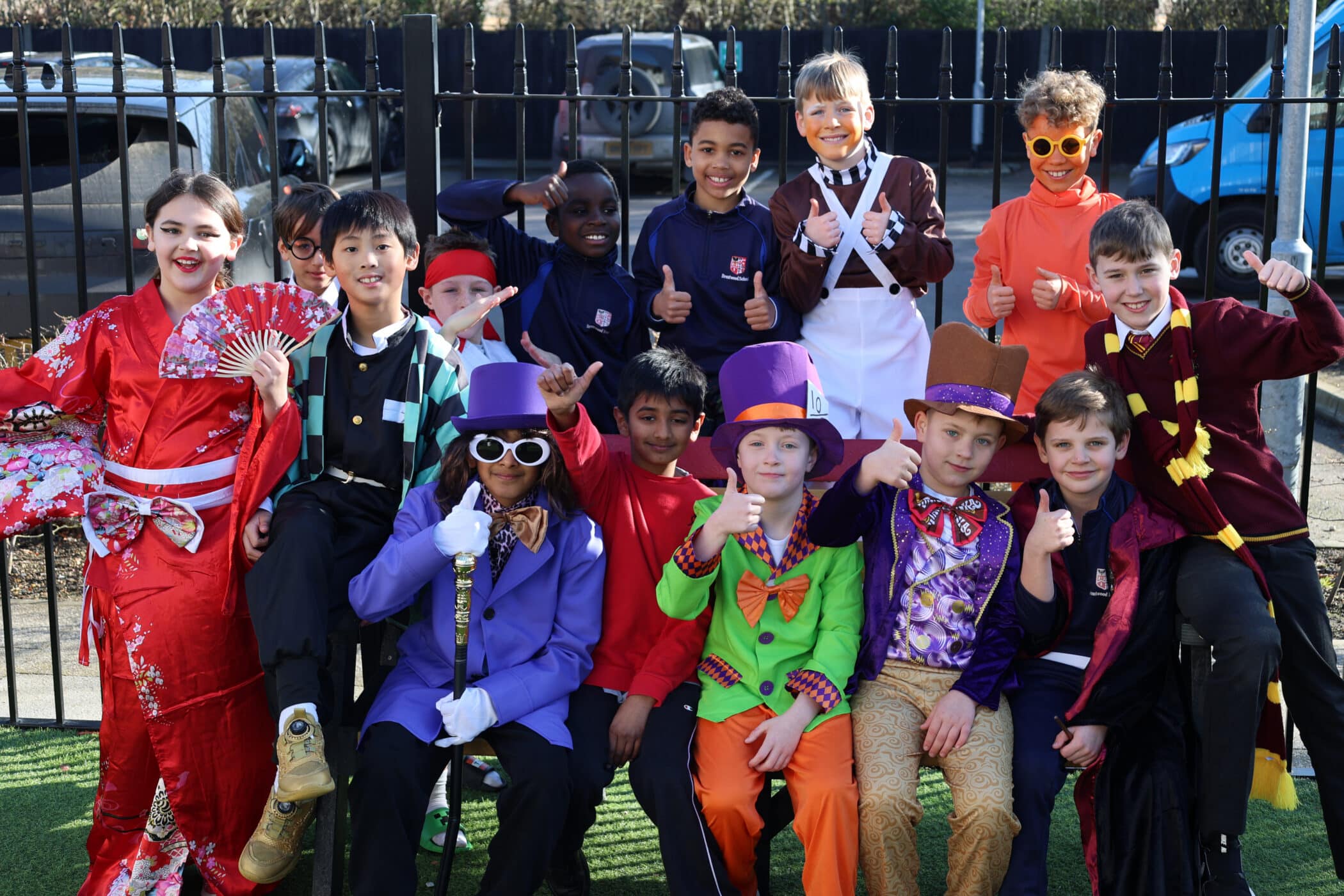 A group of children in colourful costumes, including a kimono, purple hats, and character outfits, pose outdoors in front of a black fence, smiling and giving thumbs up.