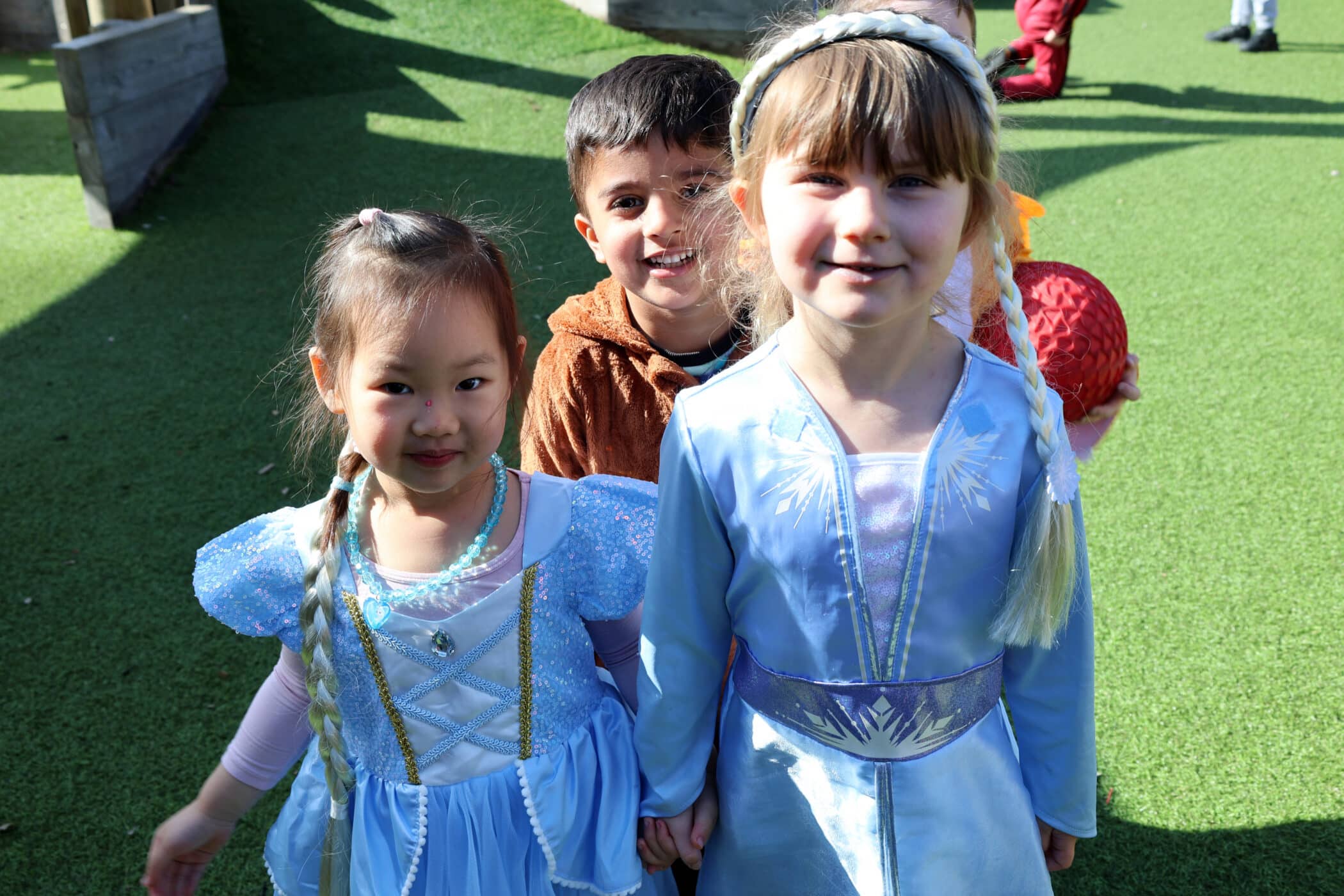 Three young children stand outdoors on artificial grass, smiling at the camera. Two girls wear blue princess fancy dress costumes, and a boy in brown stands behind them. Sunlight creates shadows on the ground.