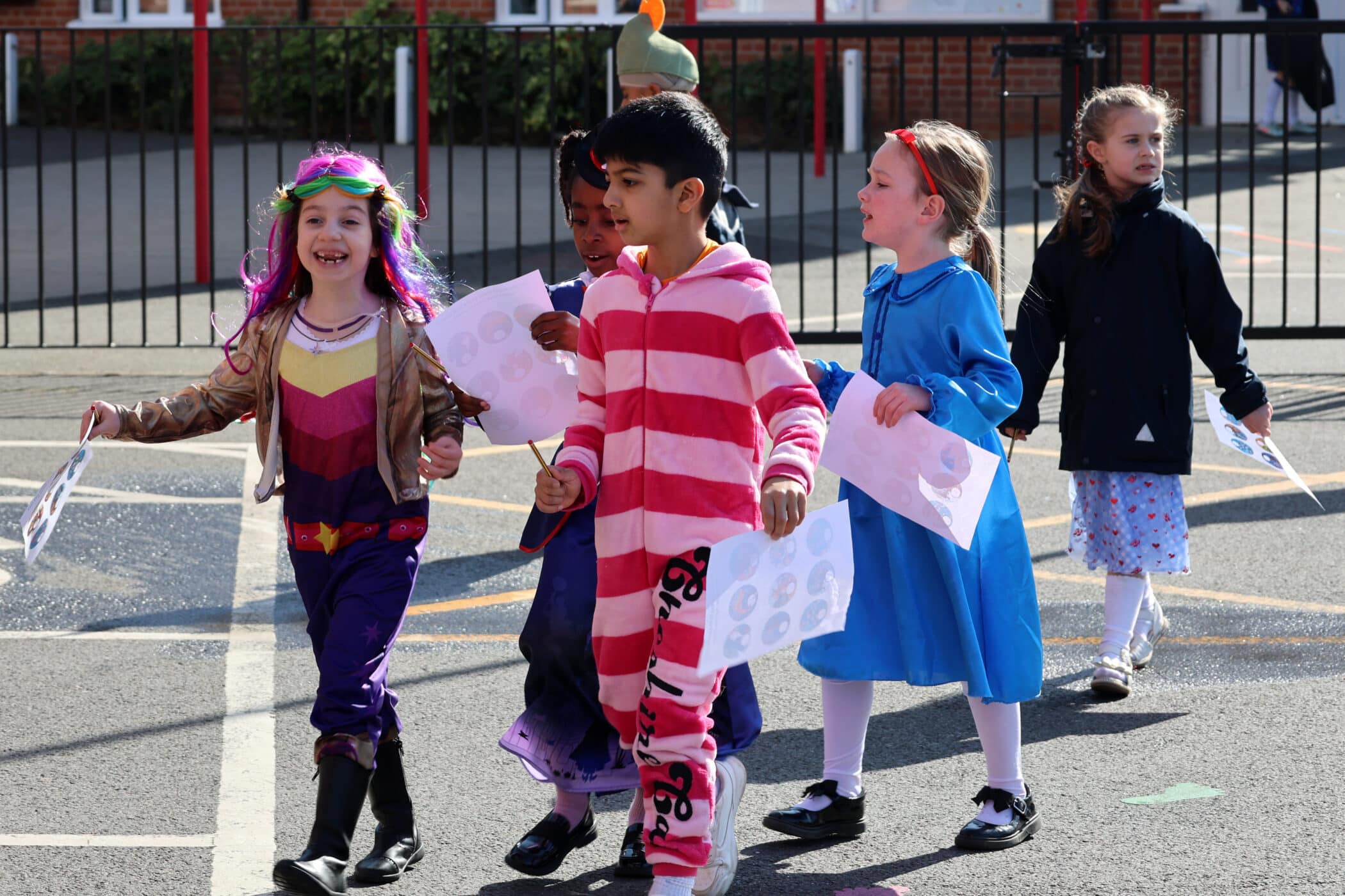 Five children in colourful costumes walk together outside, holding papers. They appear to be participating in a school event or parade, with one dressed as Wonder Woman and another in a striped pink onesie.