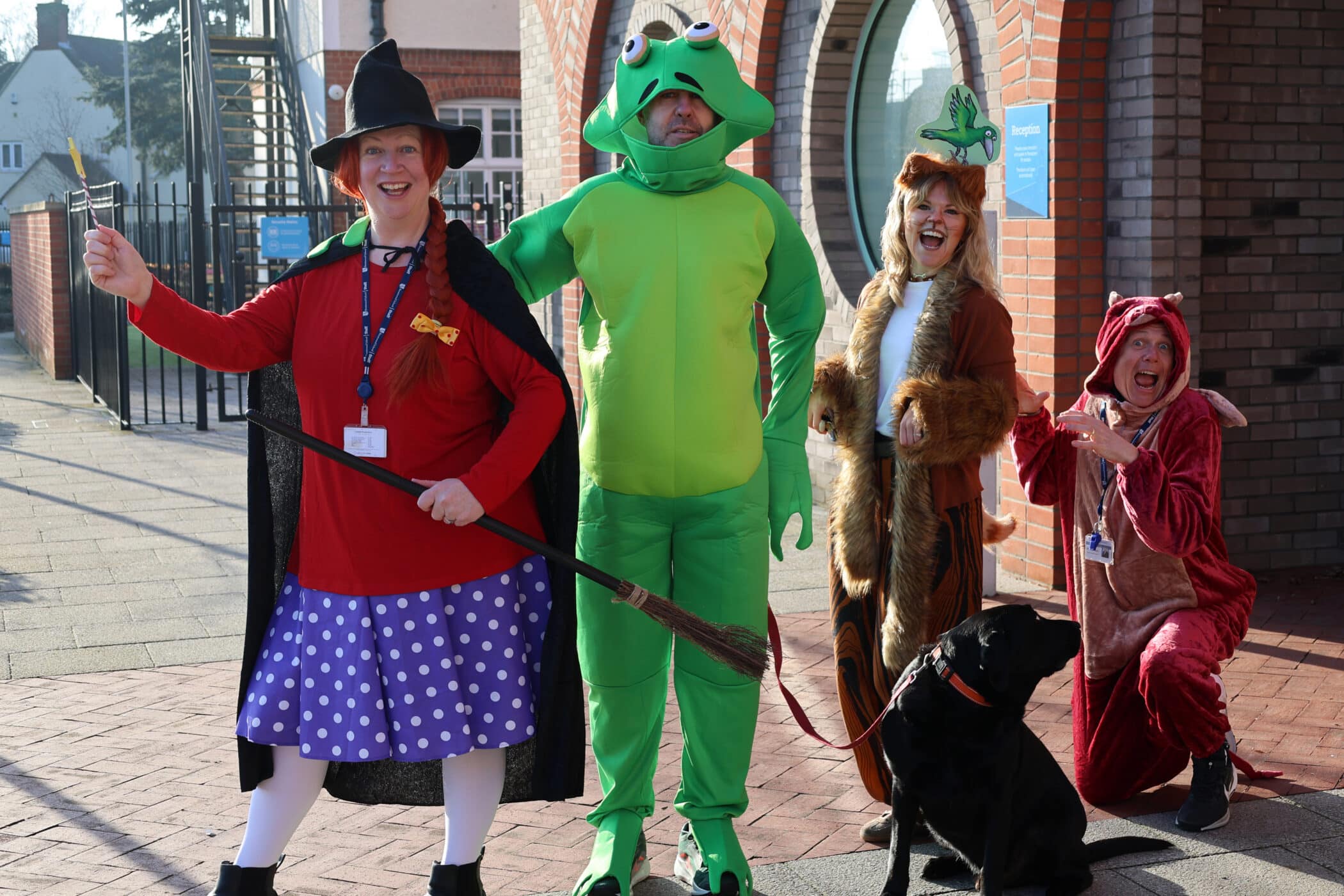 Four adults in colourful animal and witch costumes pose happily outside a brick building. One holds a broom, another wears a frog outfit, one is dressed as a dog with a black dog, and the last is in a dragon costume.