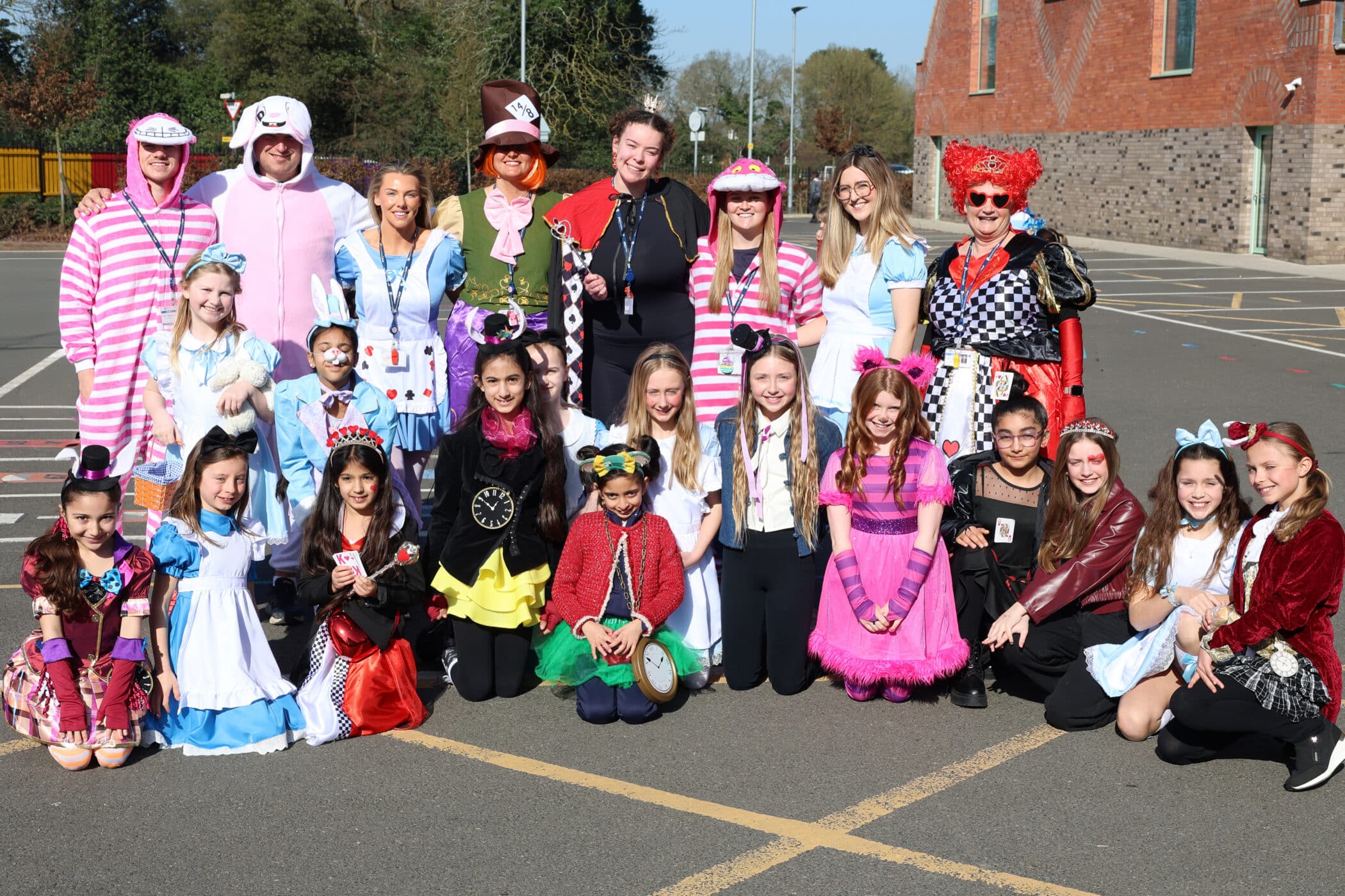 A group of children and adults in colourful costumes pose outdoors, many dressed as characters from Alice in Wonderland. They smile at the camera on a sunny day, standing in a car park with buildings in the background.