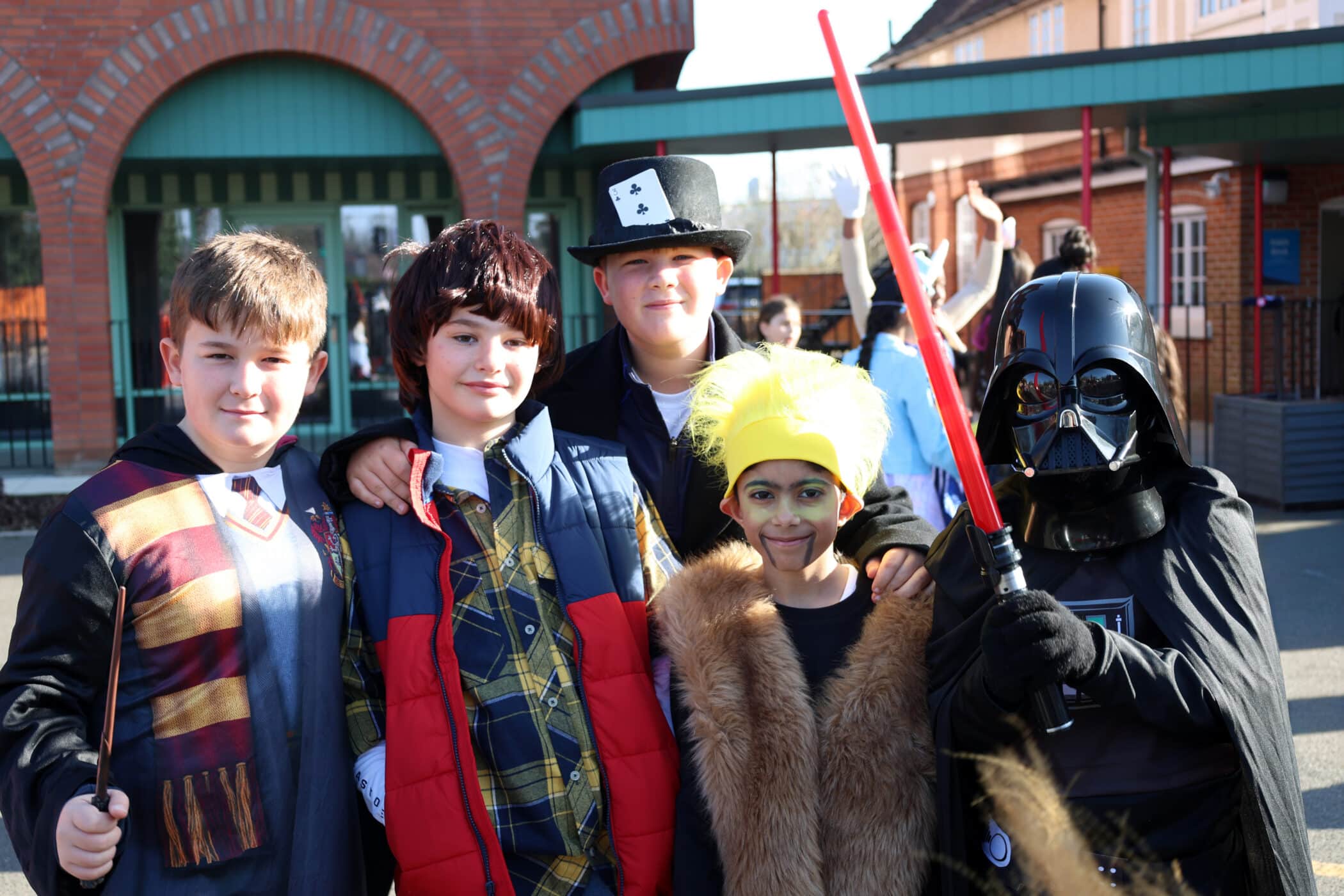 Five children in fancy dress pose outside a school. Outfits include Harry Potter, a boy in a waistcoat and checked shirt, someone in a playing card hat, a child with yellow hair and fur coat, and Darth Vader holding a red lightsabre.