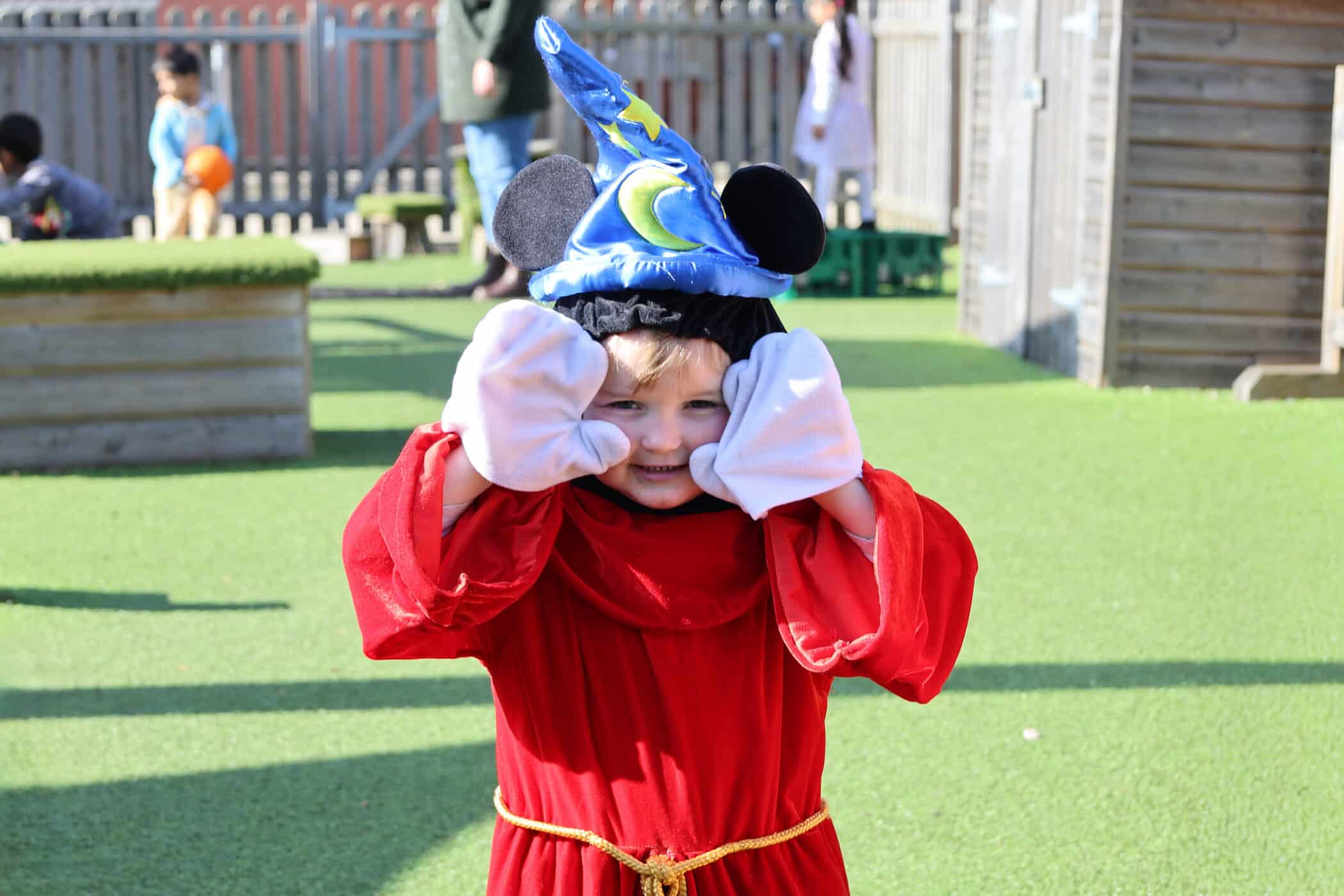 A young child dressed in a red dressing gown, large cartoon gloves, and a blue wizard hat with moon and stars plays outside on artificial grass, smiling and holding their hands to their cheeks. Other children play in the background.