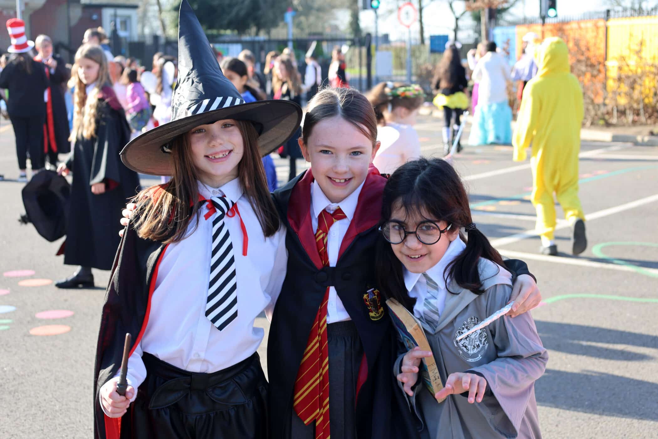 Three children dressed as Harry Potter characters stand smiling together outdoors during a fancy-dress event, surrounded by other kids in various costumes.