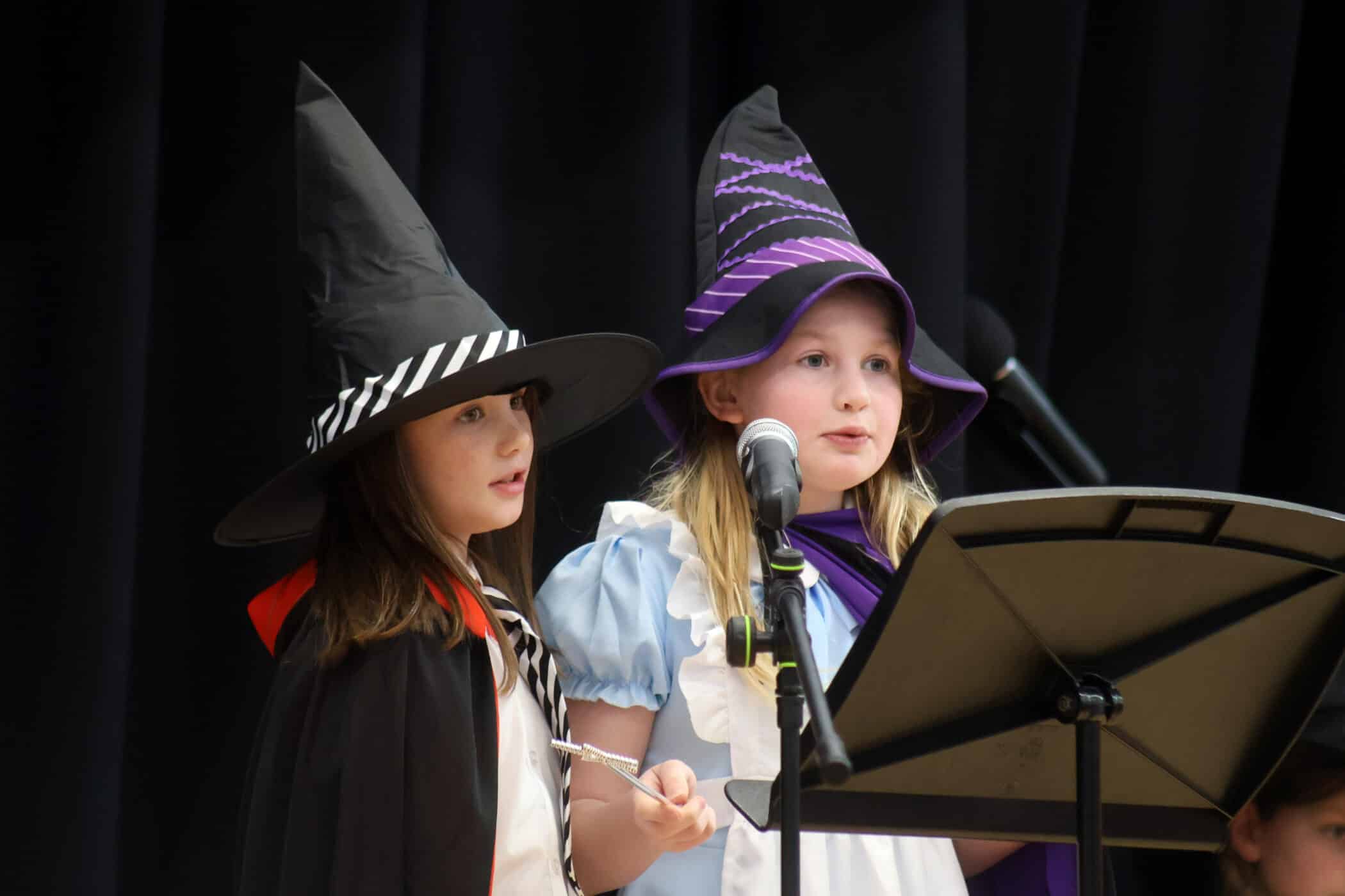 Two young girls wearing witch hats and costumes stand in front of microphones and a music stand, possibly performing or speaking on stage against a dark curtain backdrop.