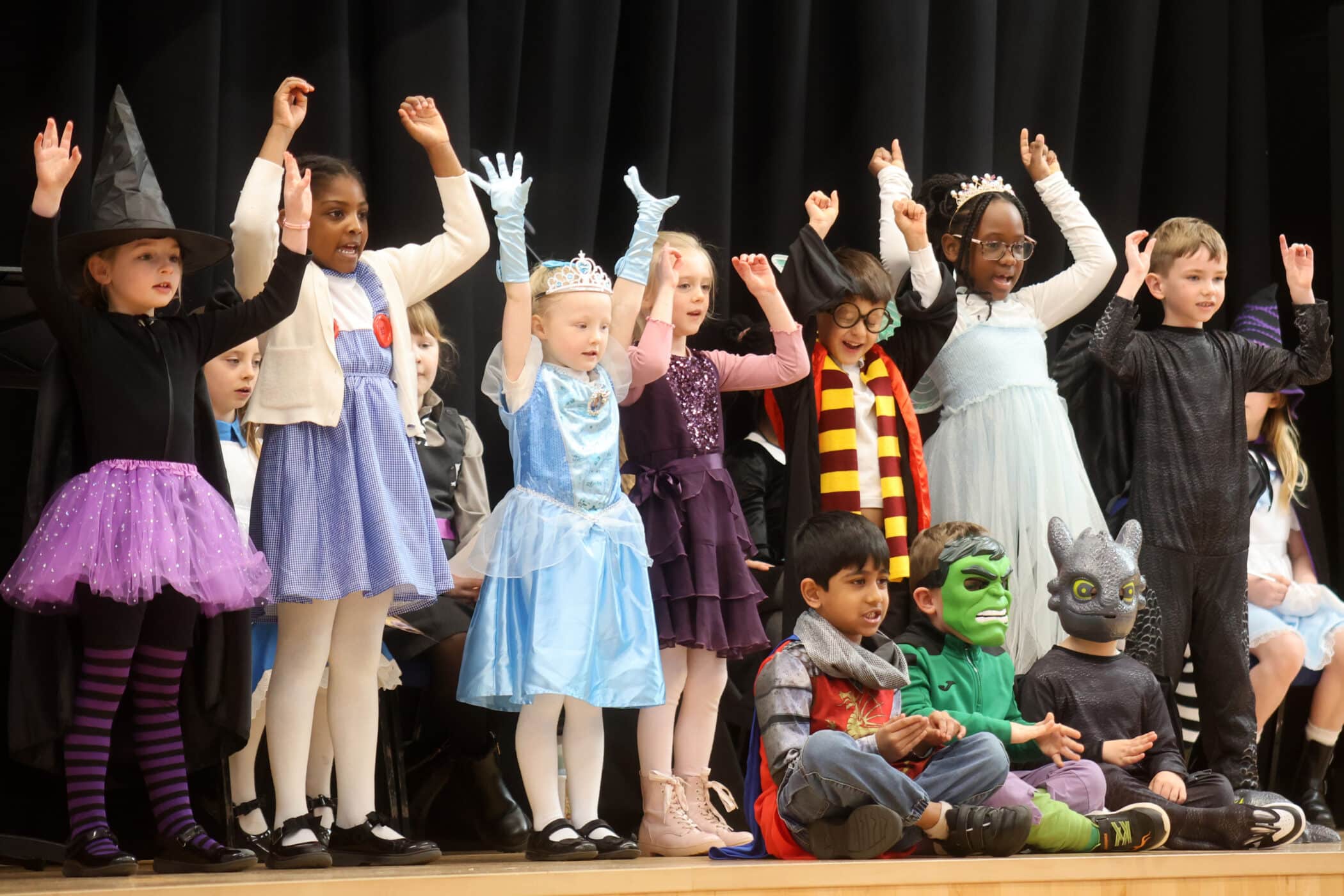 A group of children in colourful costumes, including a witch, princess, superhero, and dragon, sit and stand on a stage with their arms raised, performing in front of a black curtain.