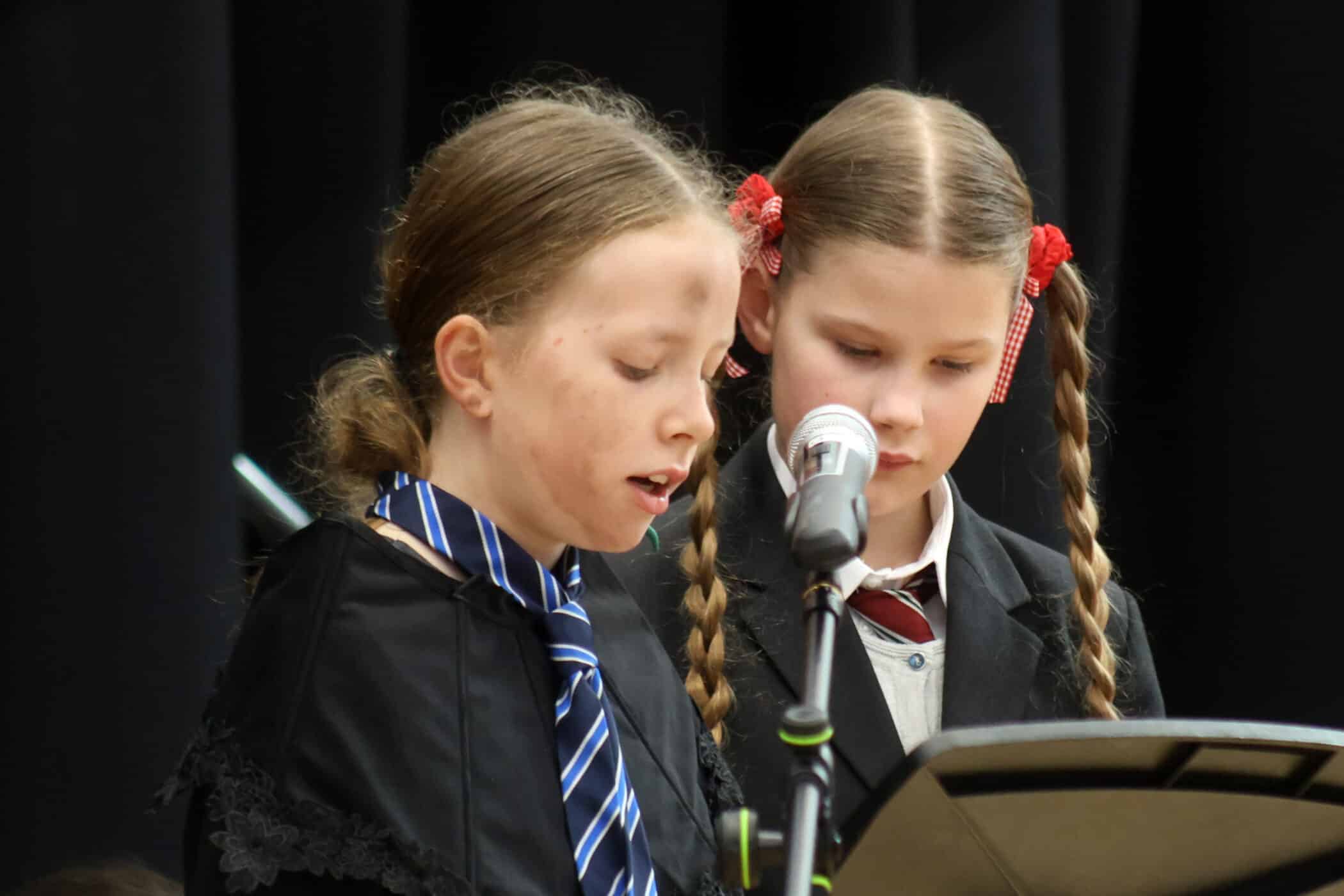 Two young girls with plaited hair, wearing fancy dress and ties, stand close together and read from a music stand while one speaks into a microphone on a stage with a dark background.
