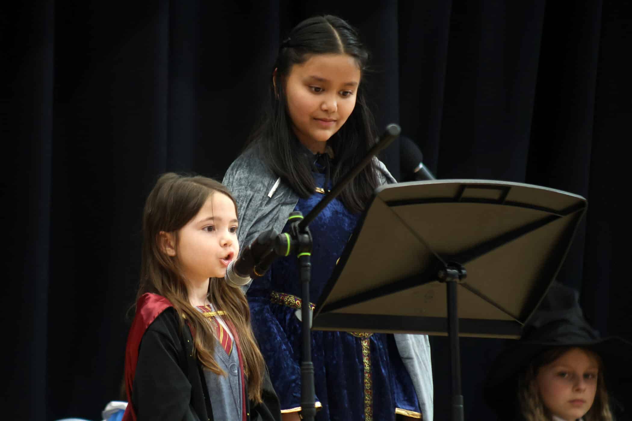 Two young girls in fancy dress stand at a music stand on stage, one speaking or singing into a microphone while the other looks on, with a dark curtain as the background. Another child in a hat is partially visible to the right.