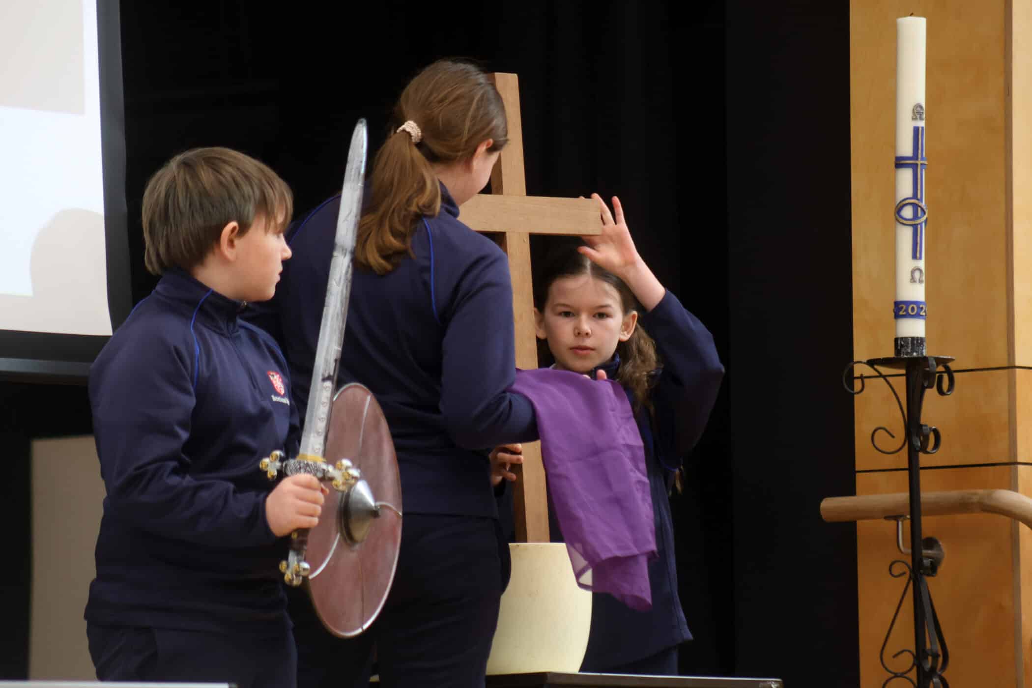 Three children in school uniforms perform on stage. One holds a toy sword and shield, while another drapes a purple cloth over a wooden cross. A large candle marked “2023” stands nearby.