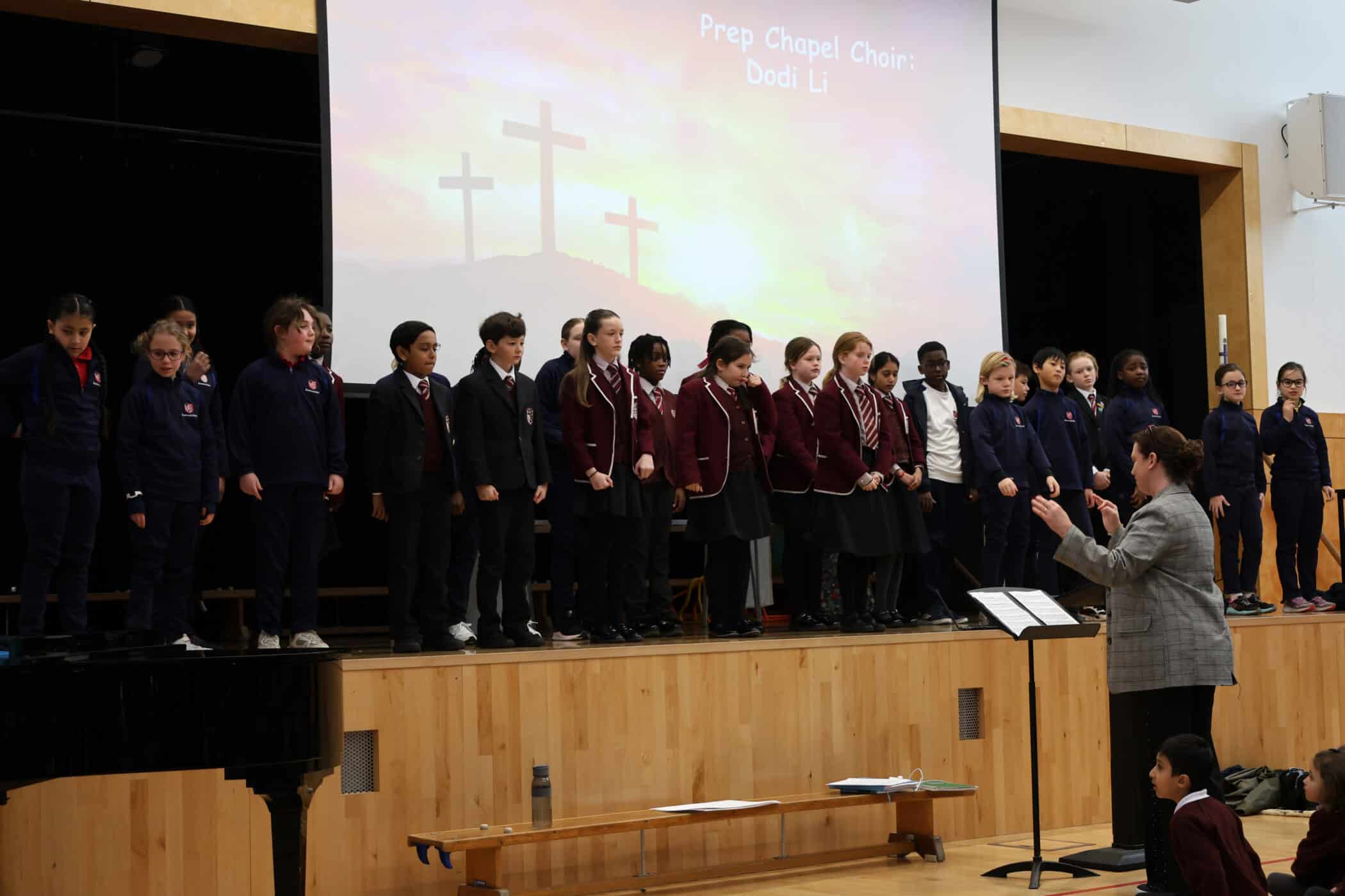 A group of students in uniforms stand on stage singing, with a conductor facing them. Behind them is a screen showing three crosses on a hill and the text “Prep Chapel Choir: Dadi Li”.