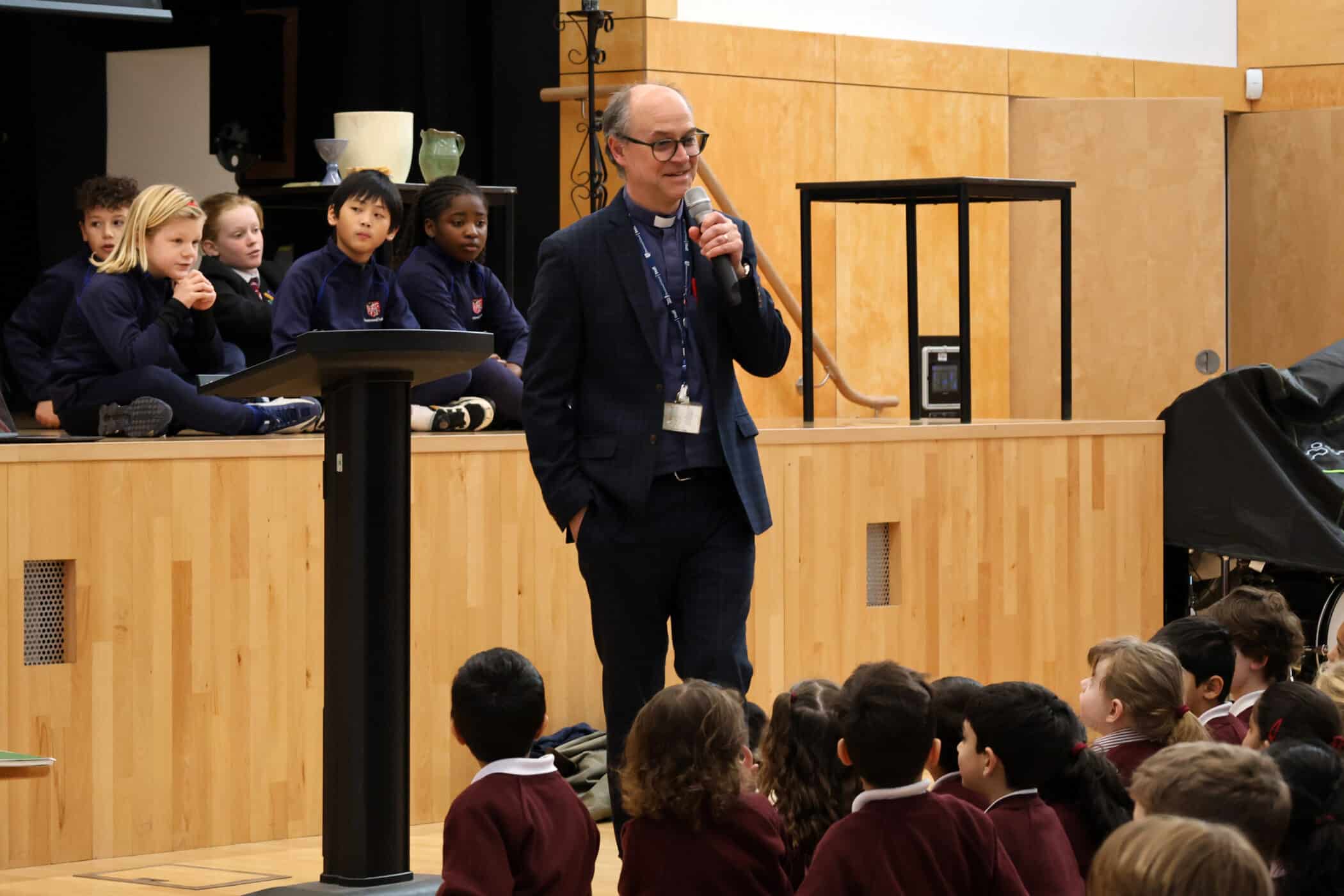 A man in a suit speaks into a microphone in a school hall, standing near a lectern. Children in uniforms sit on the stage behind him and on the floor in front, listening attentively.