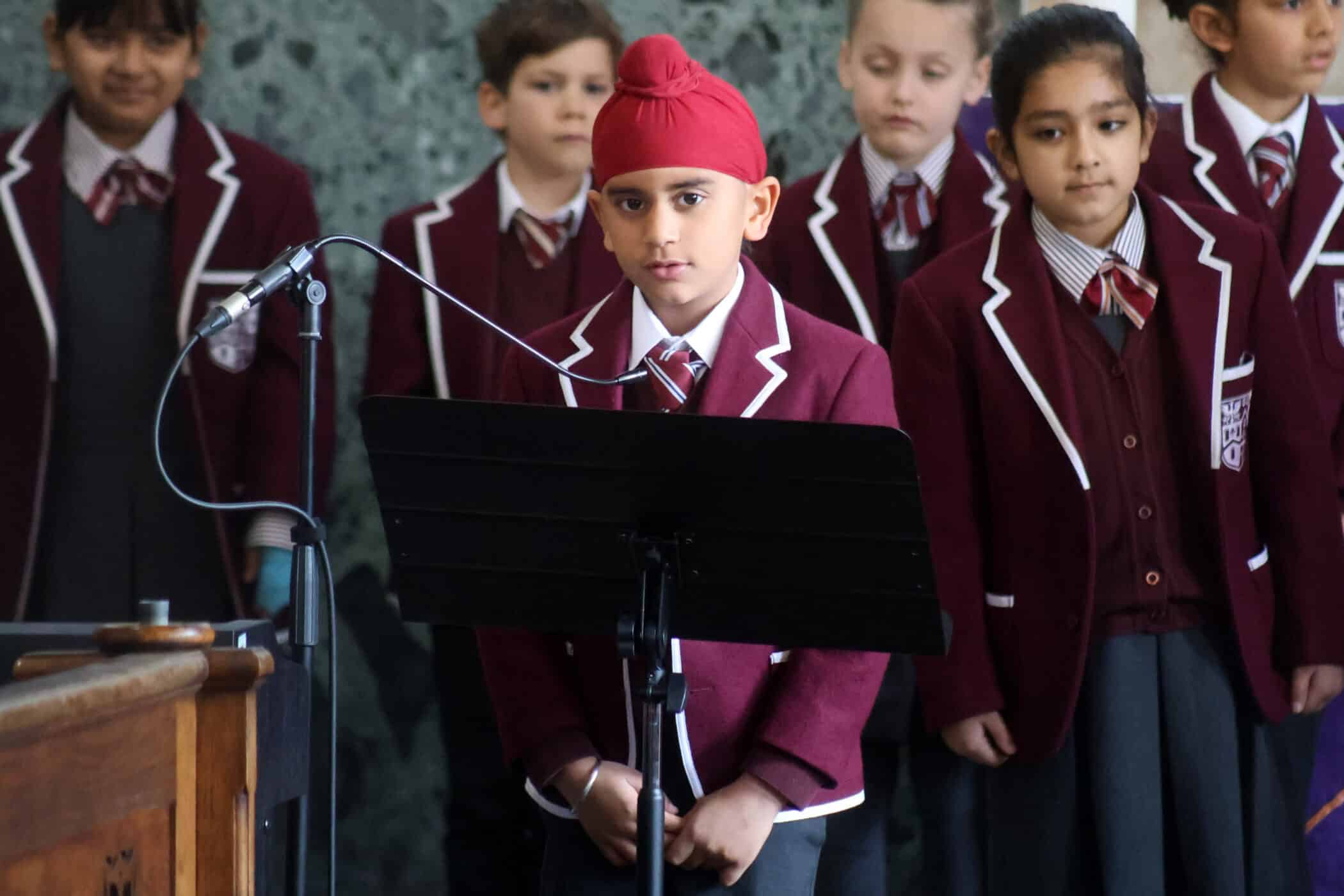 A group of schoolchildren in maroon blazers and striped ties stand together. A boy wearing a red turban stands at a microphone and music stand, appearing to prepare to speak or sing.