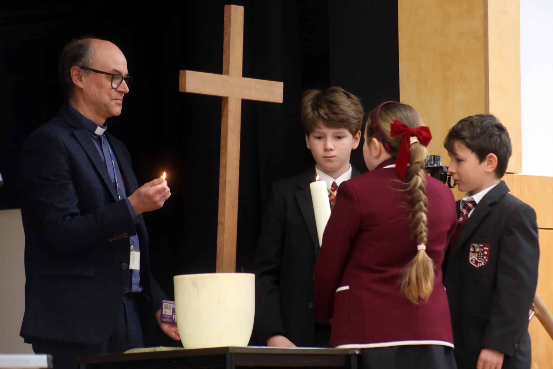 A man in glasses holds a lit match near a candle on a table beside a wooden cross, while three schoolchildren in uniforms stand nearby, two facing away and one facing the man.