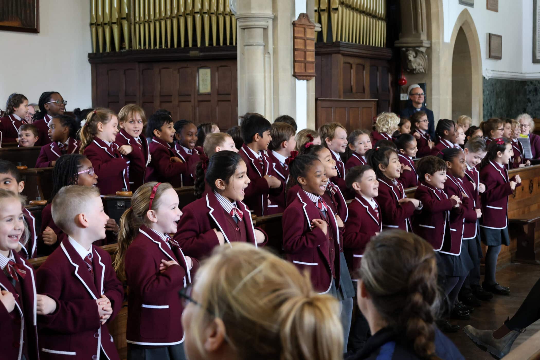 A group of young children in matching maroon blazers sing and gesture enthusiastically in a church with wooden pews and a large organ in the background. Adults are seated in the foreground, watching the performance.