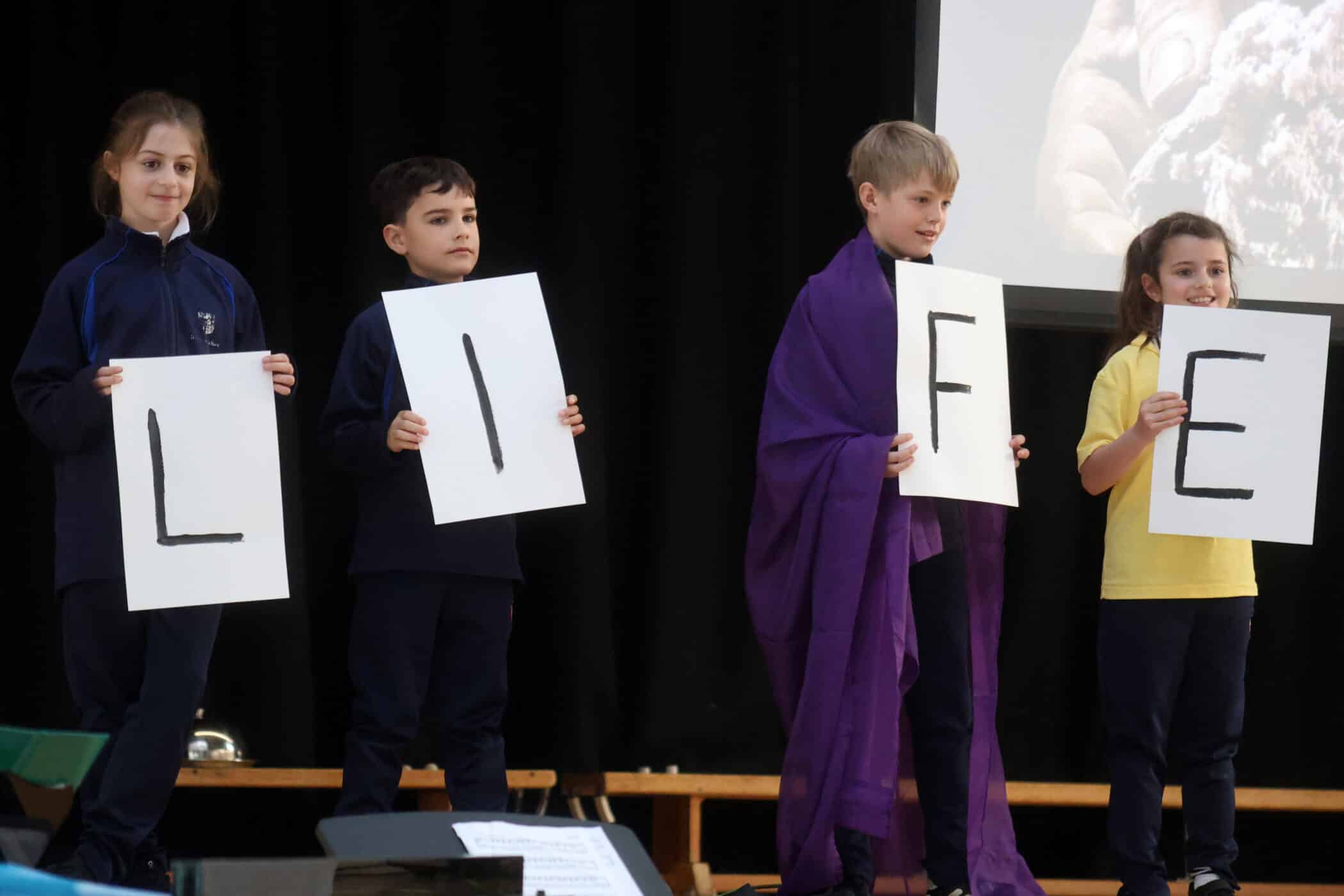 Four children stand on stage holding large white cards with the letters L, I, F, and E, spelling LIFE. The background is dark, and one child wears a purple cloth over their shoulders.