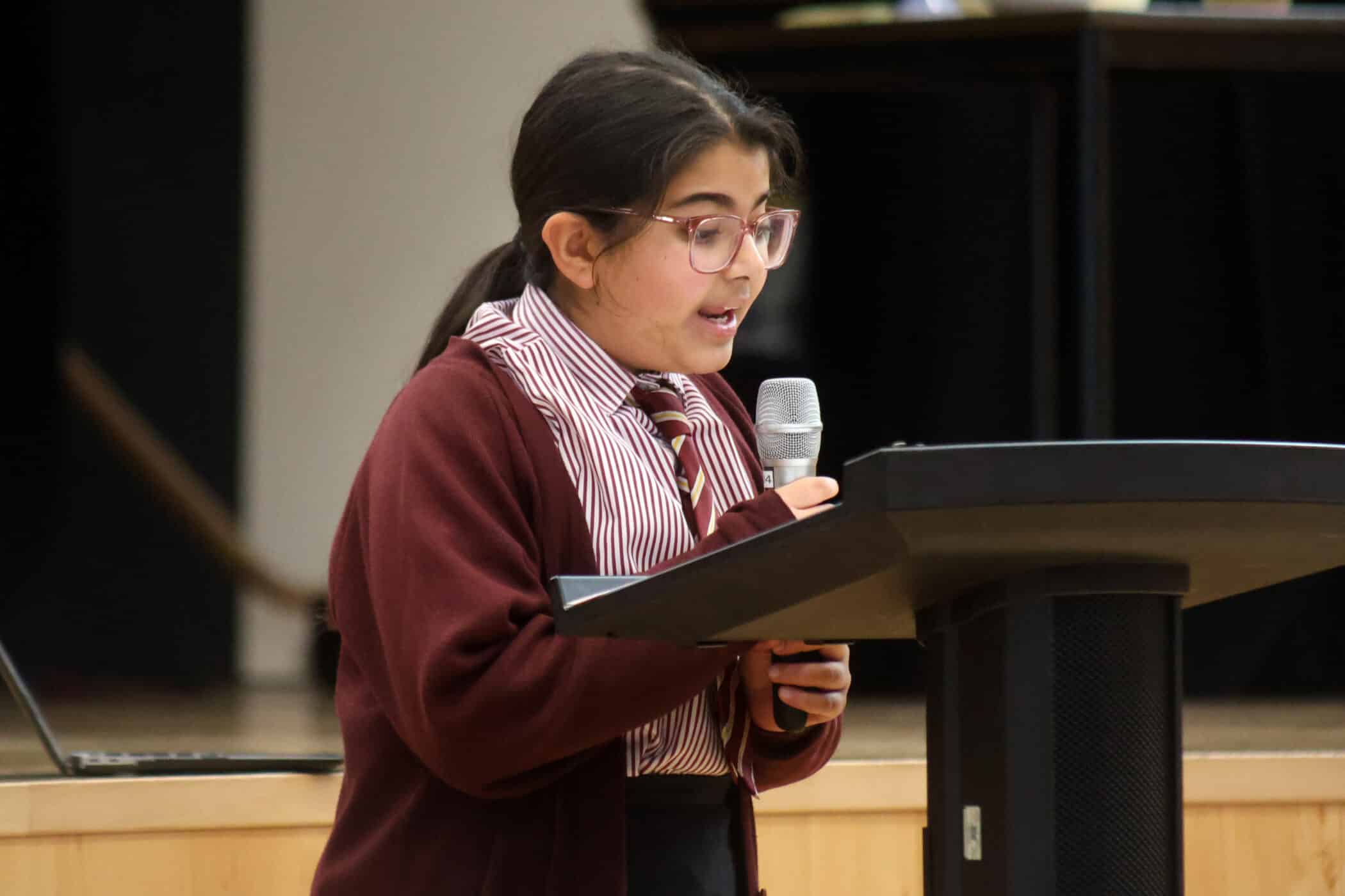 A student wearing glasses and a maroon uniform speaks into a microphone whilst standing at a lectern, possibly delivering a presentation or speech in a school setting.