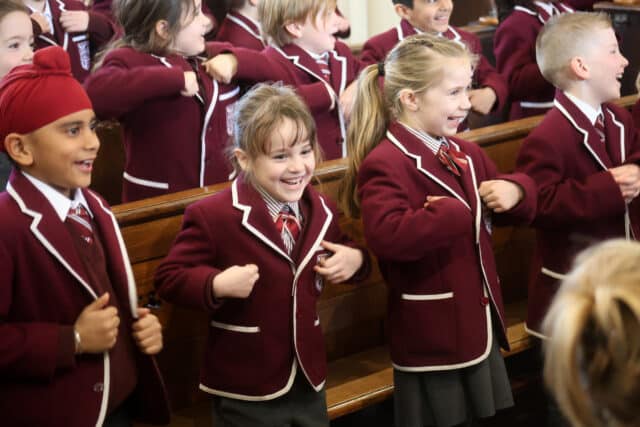 A group of young schoolchildren wearing maroon blazers and ties stand in a row, smiling and laughing whilst performing actions with their arms in what appears to be a classroom or assembly setting.