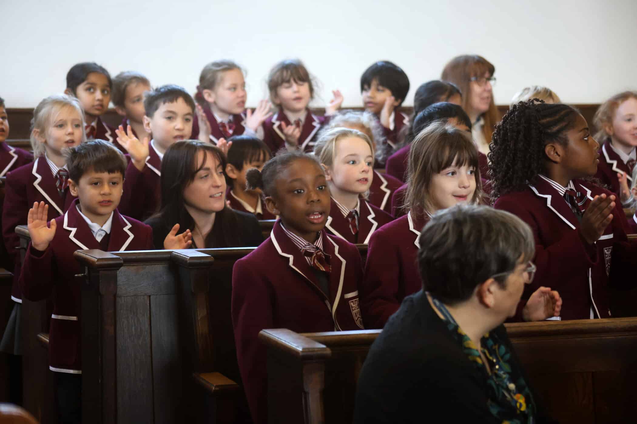 A group of young children in matching maroon school uniforms sit and raise their hands on a wooden pew, appearing attentive and engaged, with a few adults seated among them.