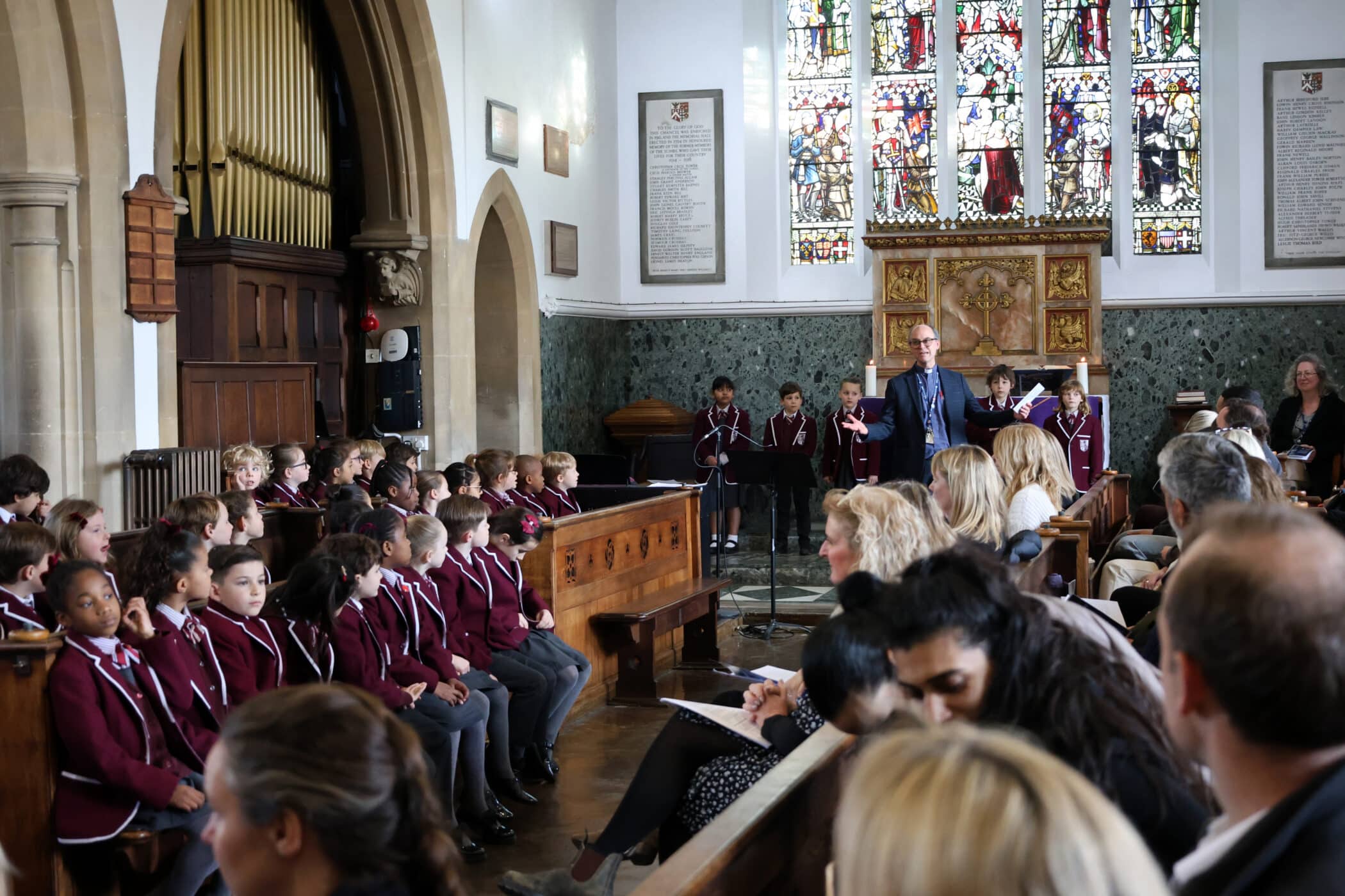 A group of children in matching maroon uniforms sit in wooden pews inside a church, with adults seated in front. A man stands at the front near a lectern, speaking, whilst others stand beside him near stained glass windows.
