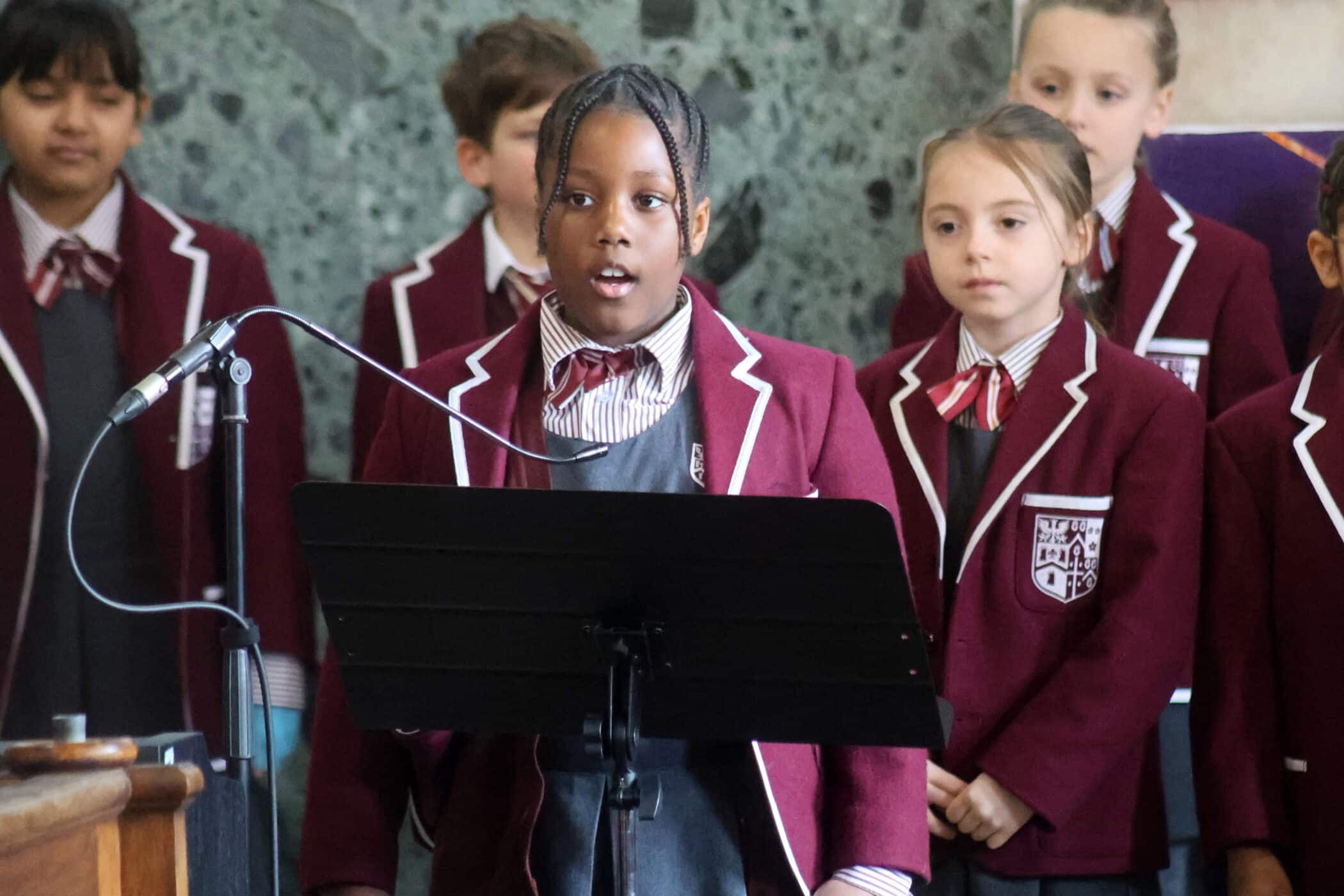 A group of schoolchildren in maroon blazers and ties sing together. One girl stands at the front, singing into a microphone with a music stand in front of her. Other children stand behind her, listening attentively.
