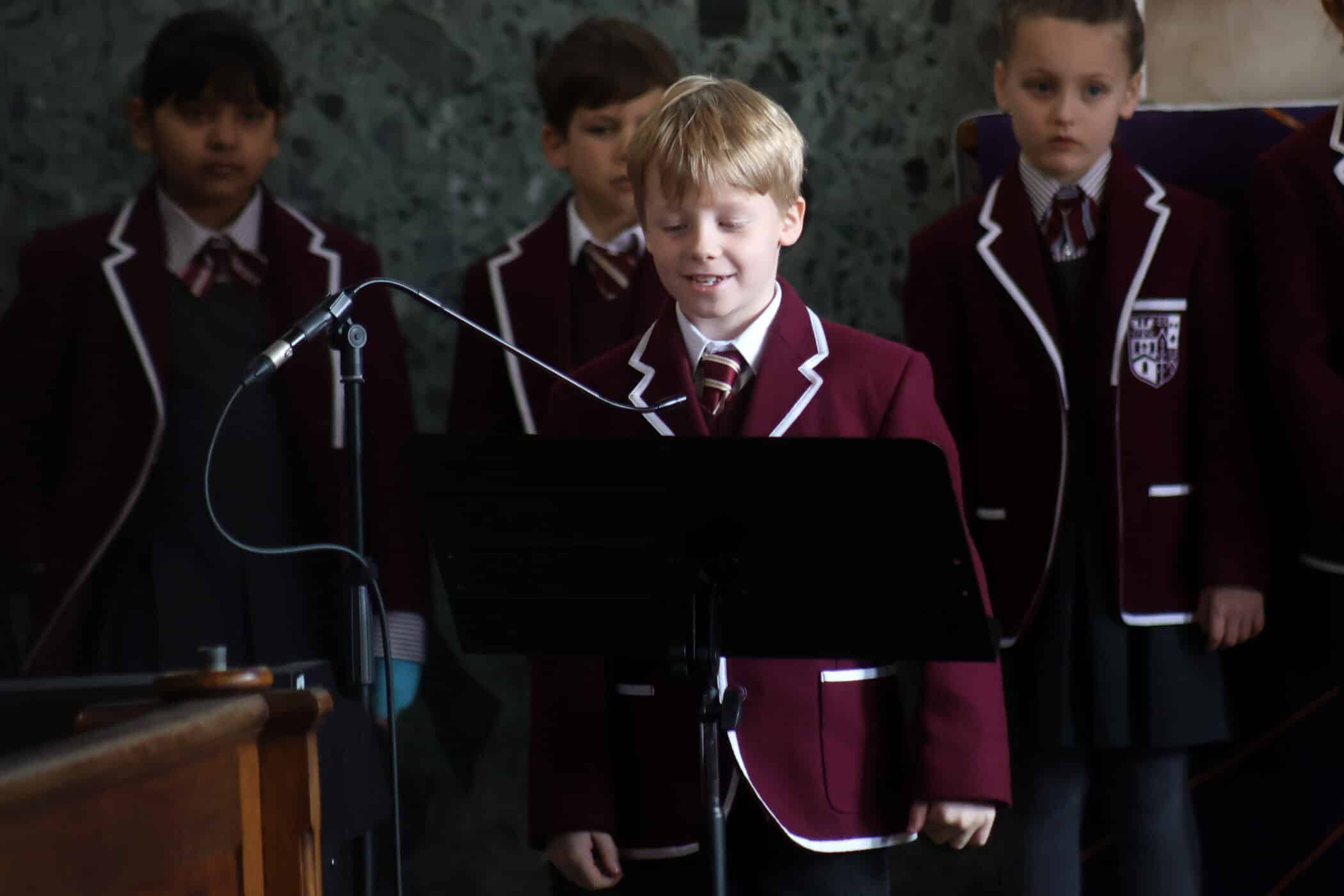 A young boy in a maroon school blazer stands at a music stand with a microphone, singing or speaking, whilst other pupils in matching uniforms stand behind him.