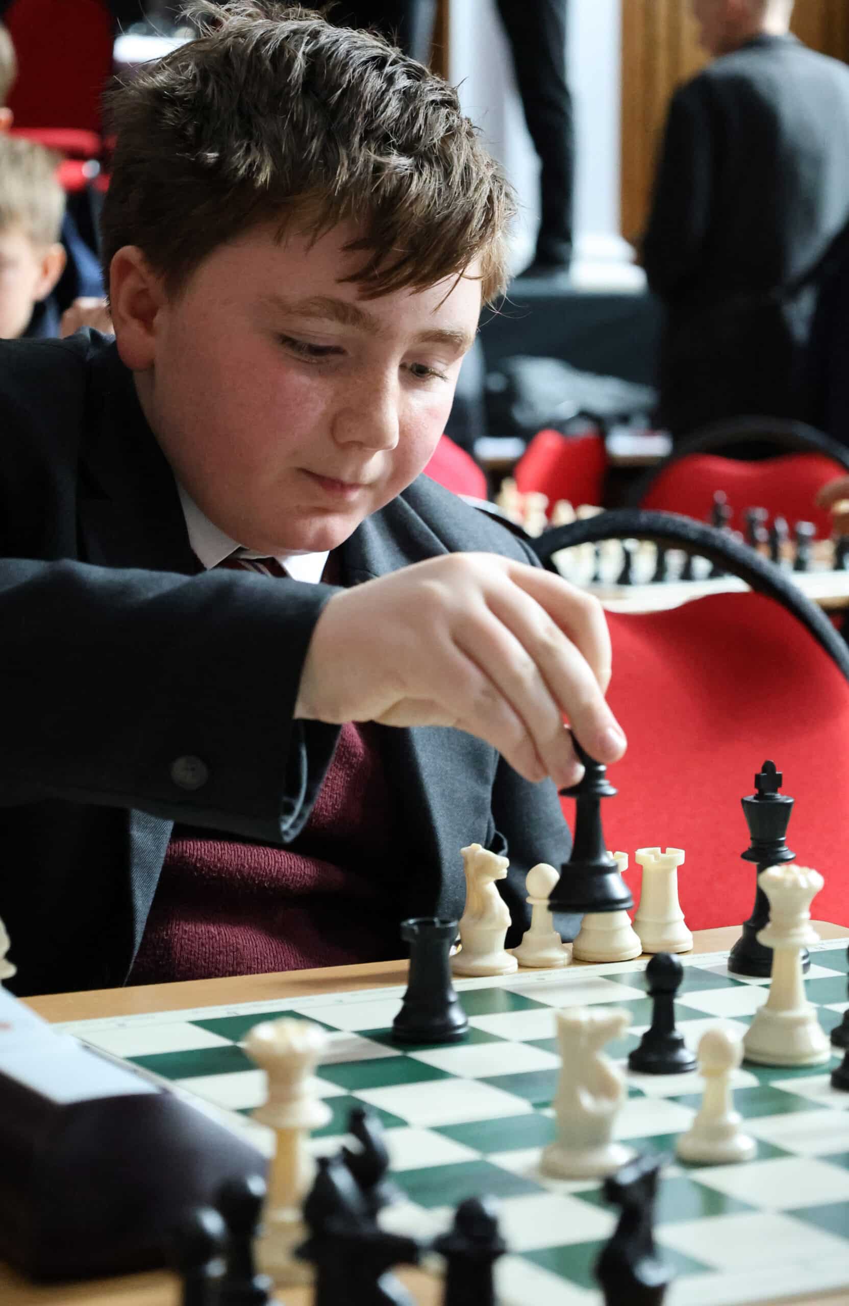 A boy in a suit concentrates as he moves a chess piece during a chess game in a tournament setting, with red chairs and other players visible in the background.