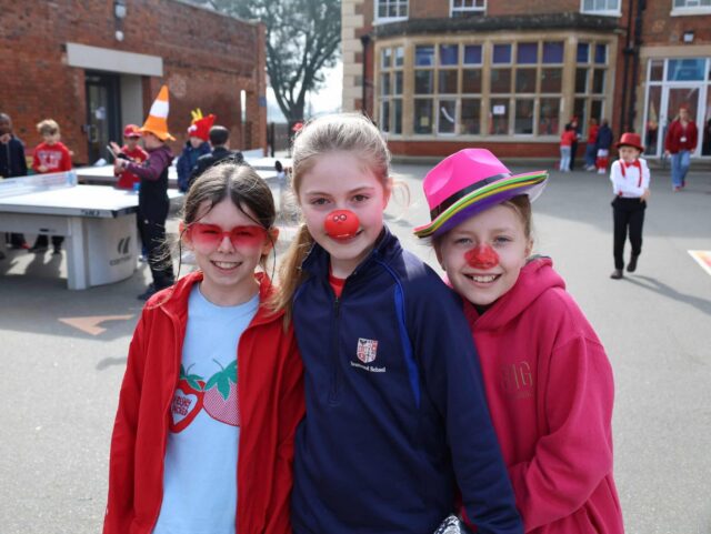 Three smiling children stand close together outside. They wear red, with two of them sporting red clown noses and fun hats or glasses. Other kids and a table tennis table are visible in the background.