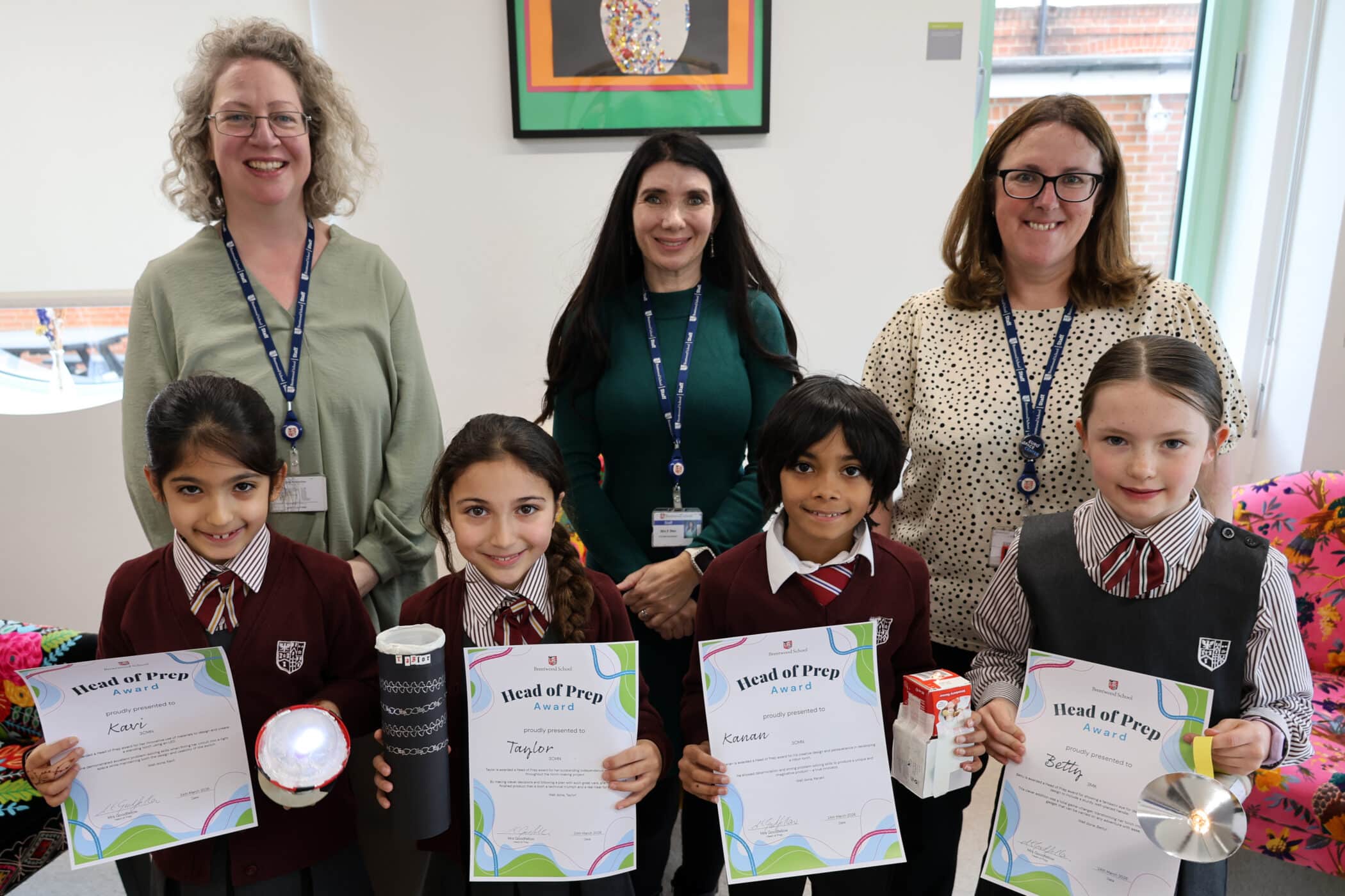 Four children in school uniforms hold certificates and projects while standing in front of three smiling adults. They are indoors, and all appear to be celebrating an achievement.