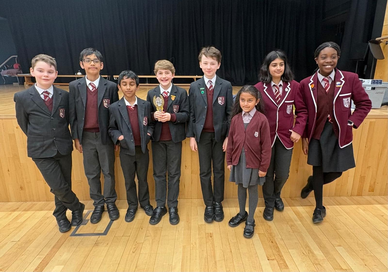 Eight pupils in matching school uniforms stand together indoors on a wooden floor, smiling at the camera. One pupil in the centre holds a trophy. A stage with a black curtain is in the background.