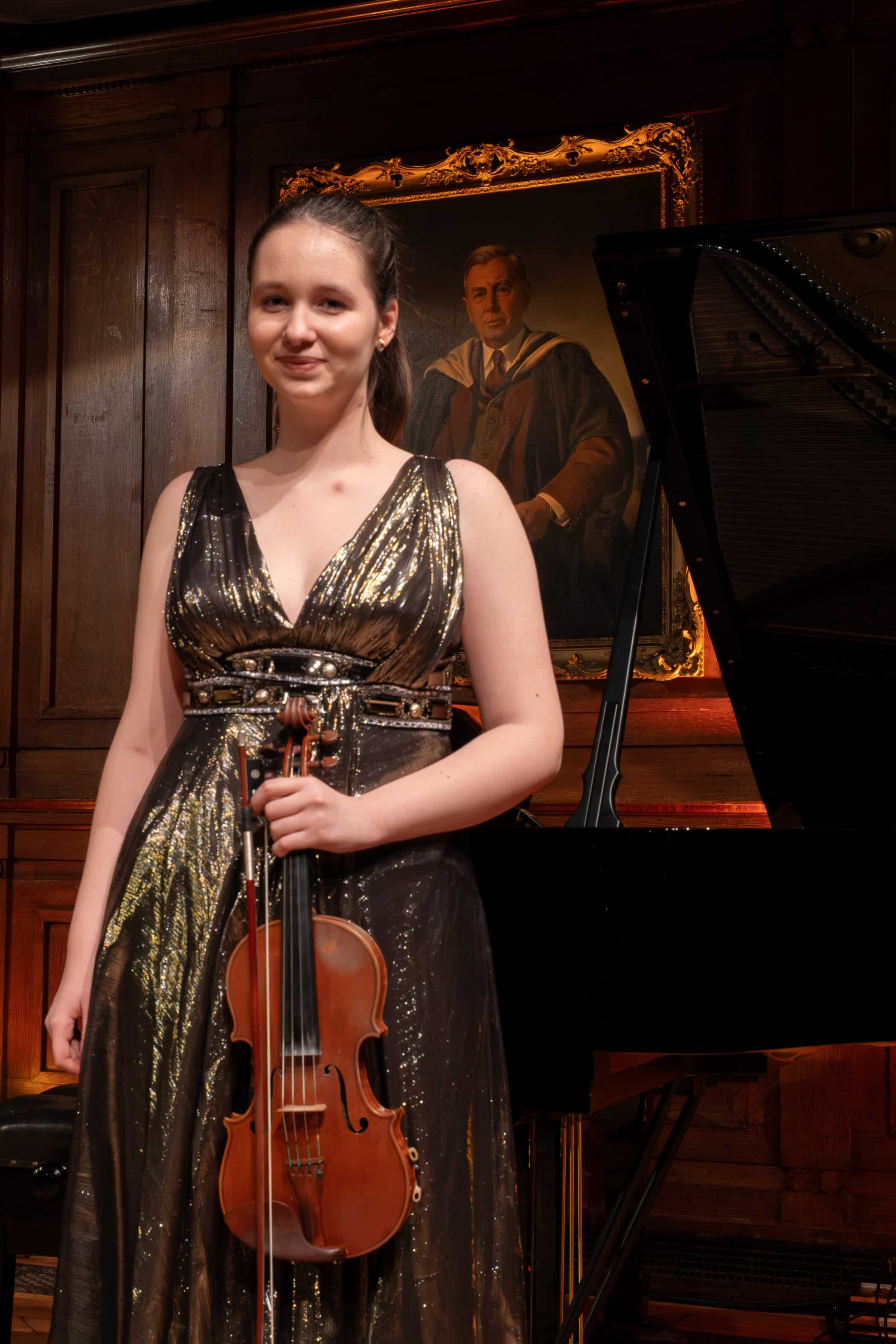 A woman in a shimmering gold and black dress stands holding a violin in front of a grand piano, at the Brentwood International Music Competition, with a portrait of a man in academic attire hanging on the wood-panelled wall behind her.