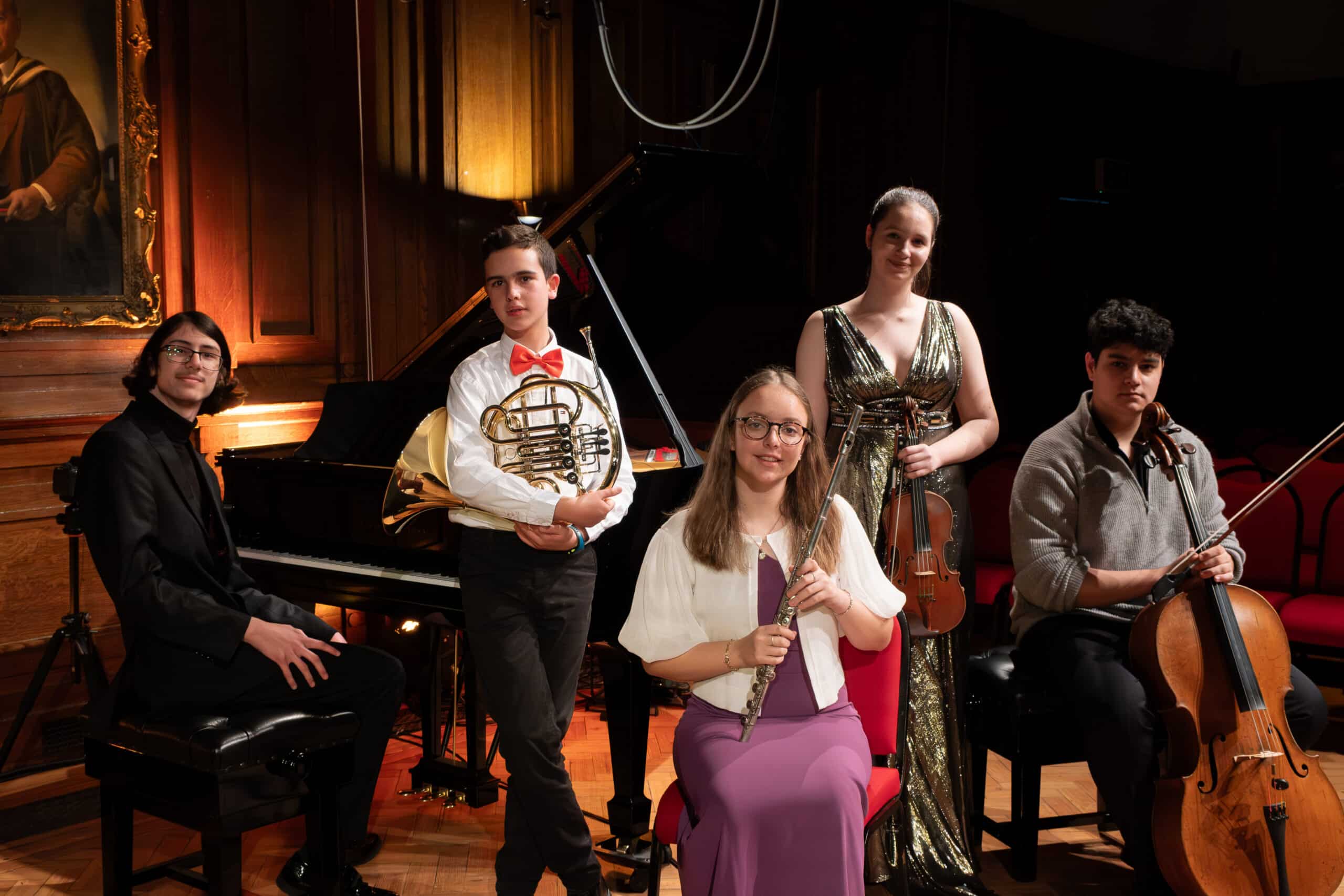 Five young musicians, finalists in the Brentwood International Music Competition, pose in a wood-panelled room with piano, French horn, violin, flute, and cello. Dressed in formal concert attire, they are poised for an impressive performance.