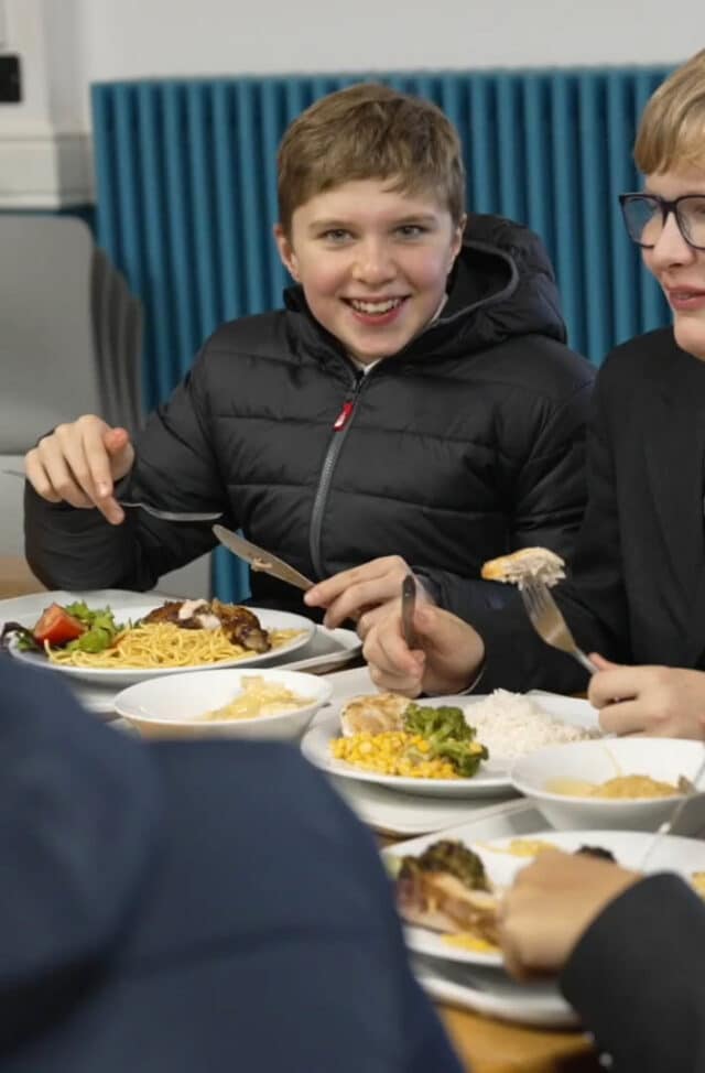 A smiling boy in a black jacket sits at a table with others at Brentwood School, enjoying a meal that includes rice, sweetcorn, salad, and noodles. He is holding a fork and knife and appears to be in a cheerful mood.