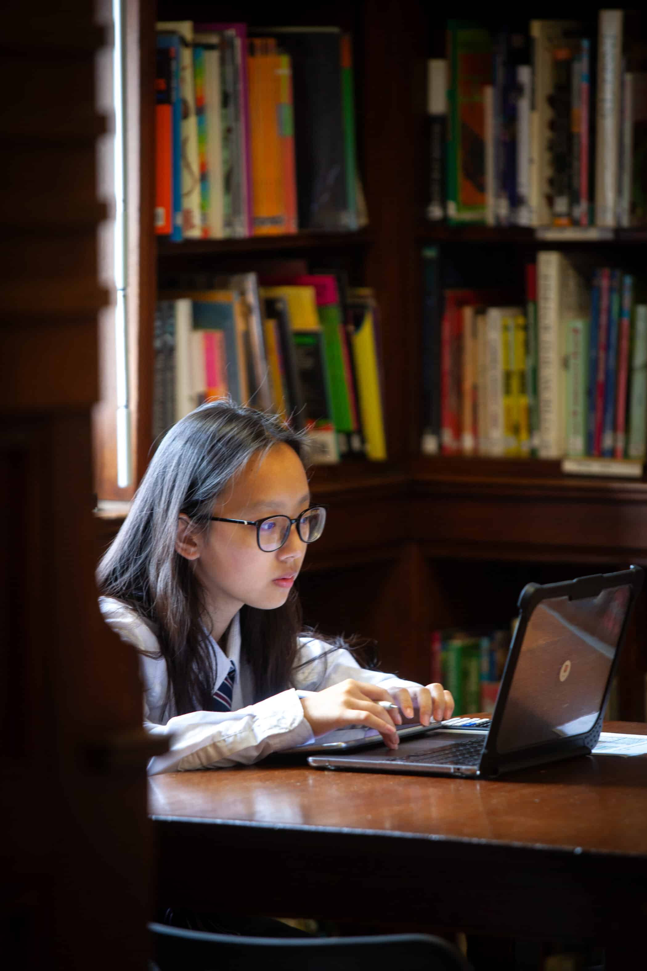 A young person with long hair and glasses sits at a wooden table in a library, focused on using a laptop, perhaps researching pastoral support during GCSE years. Bookshelves filled with colourful books are visible in the background.