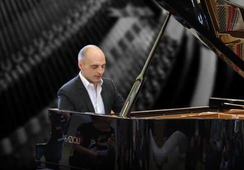 A man in a suit plays a grand piano with the lid open. The background is blurred, featuring abstract, black and white patterns that emphasise the musician’s focus on the piano.