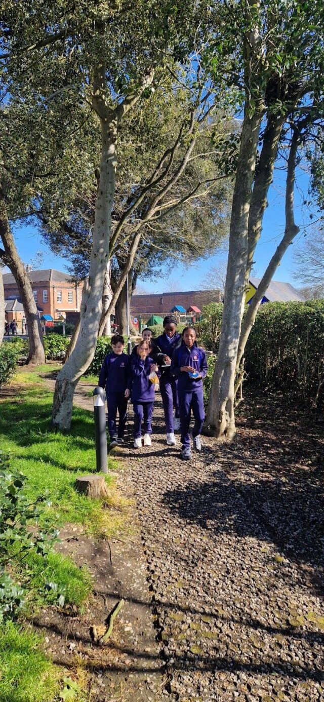 A group of five children in navy blue uniforms enjoy Walking Wednesday, strolling under tall trees on a sunny day as they follow a gravel path through a green park with houses visible in the background.