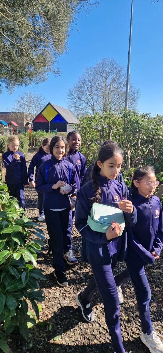 On a sunny Walking Wednesday, schoolchildren in navy blue uniforms stroll outdoors along a garden path, surrounded by green bushes and a colourful building in the background.