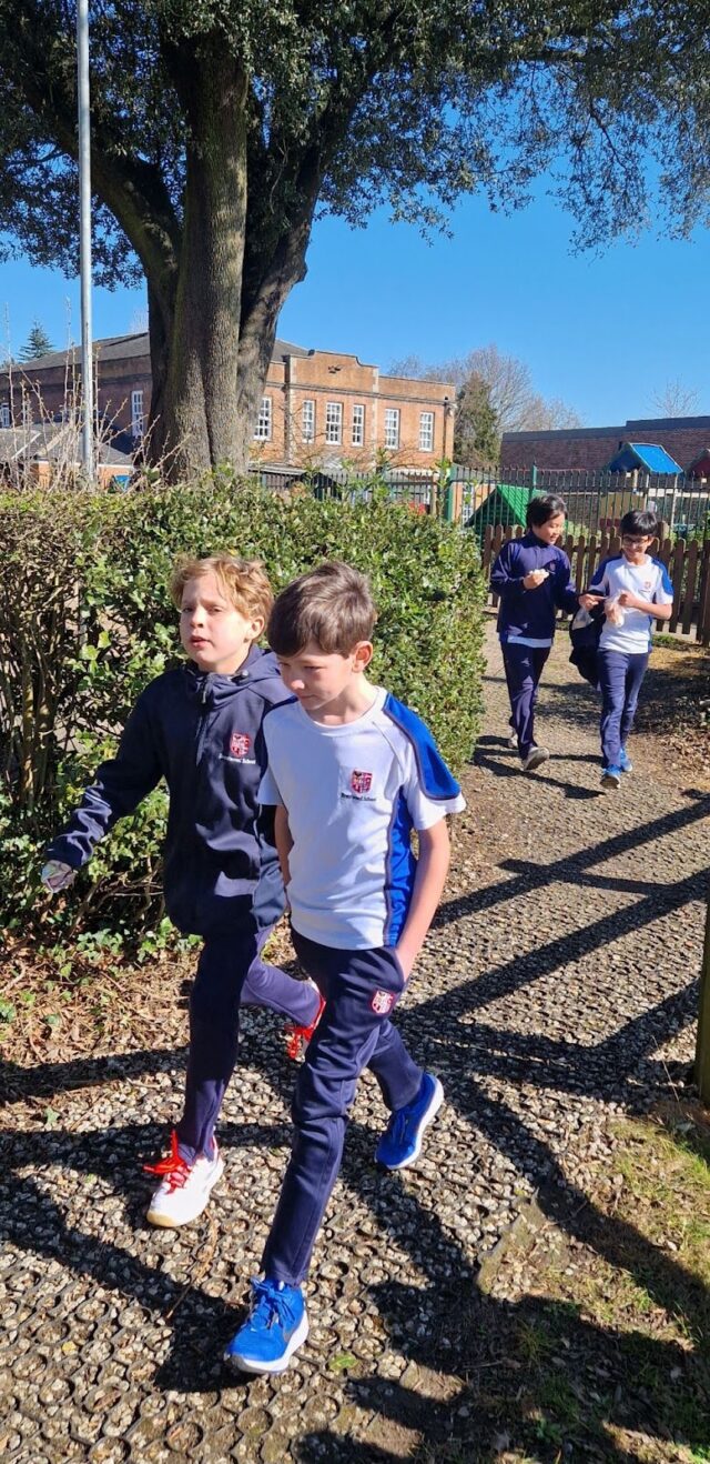 Four children in school uniforms are jogging on a gravel path outside on a sunny Walking Wednesday, with bushes on one side and a school building in the background.