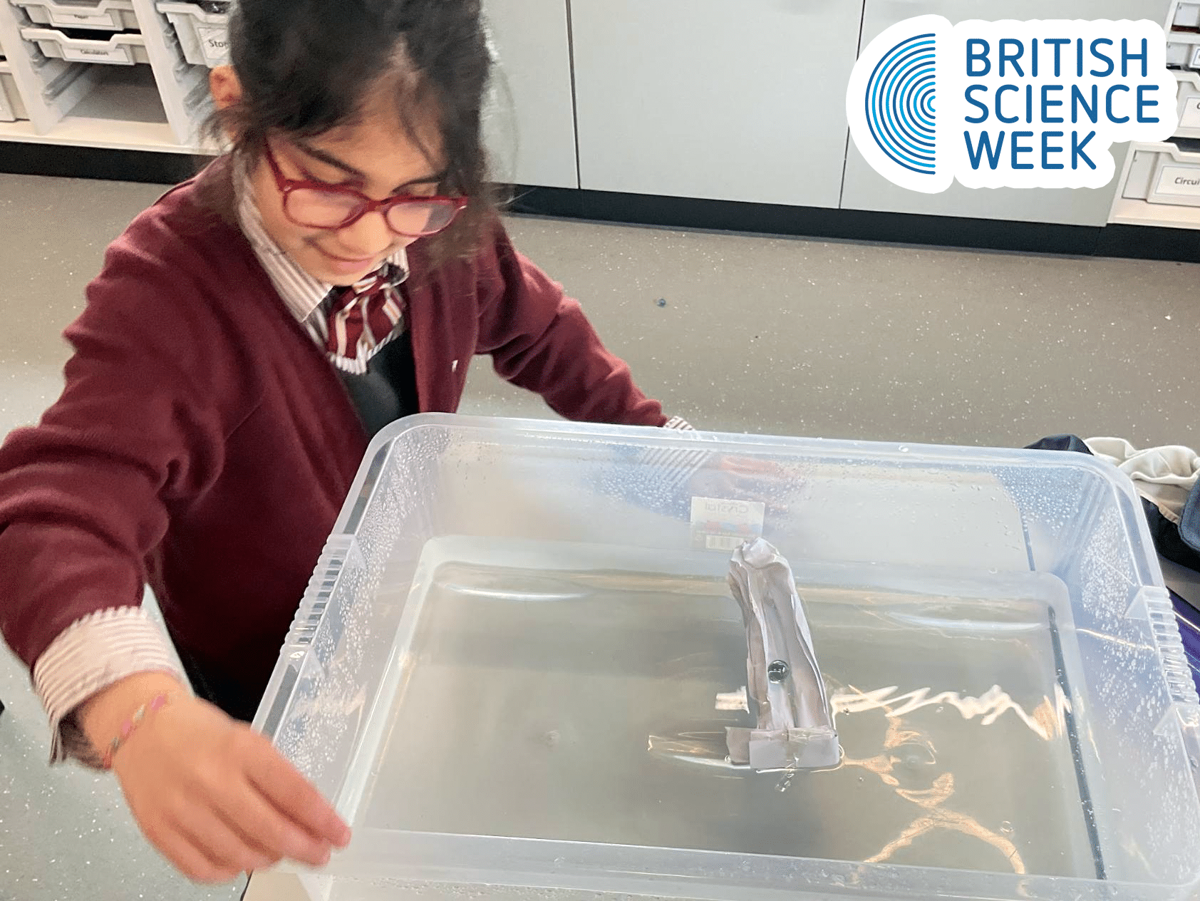 A child wearing red glasses and a maroon jumper places a small boat with a mast into a clear plastic container filled with water. The British Science Week logo appears in the top right corner.