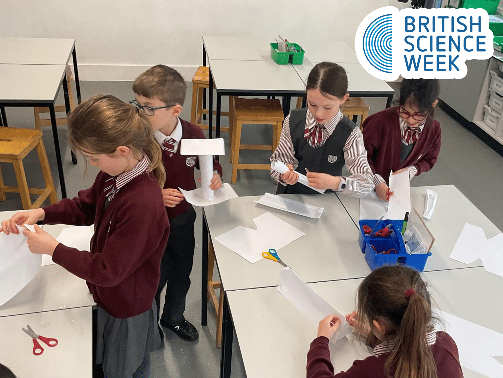 A group of schoolchildren in uniforms work on paper projects at tables during British Science Week. Scissors, paper, and other supplies are on the tables. The British Science Week logo appears in the top right corner.