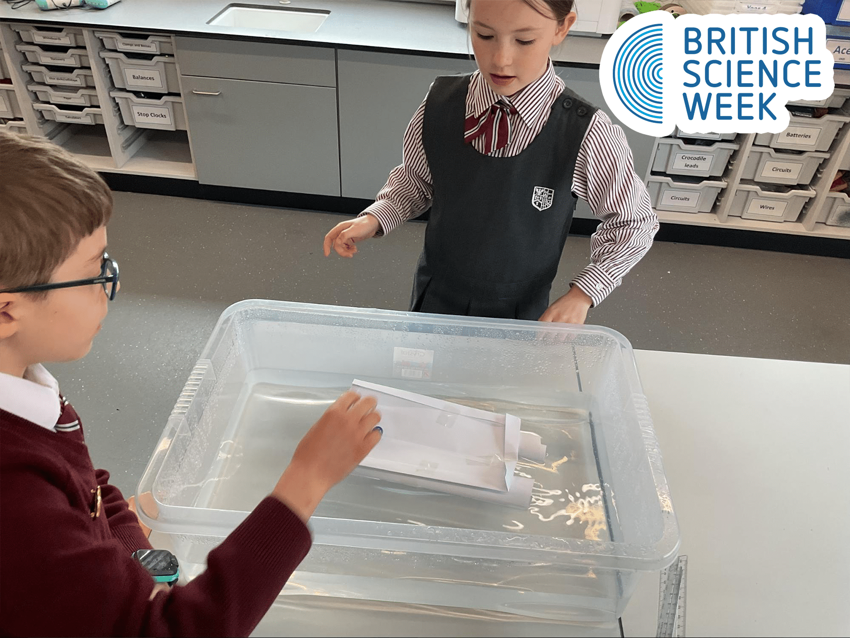 Two schoolchildren in uniforms float a paper boat in a clear plastic tub of water during a science activity. A British Science Week logo appears in the top right corner.