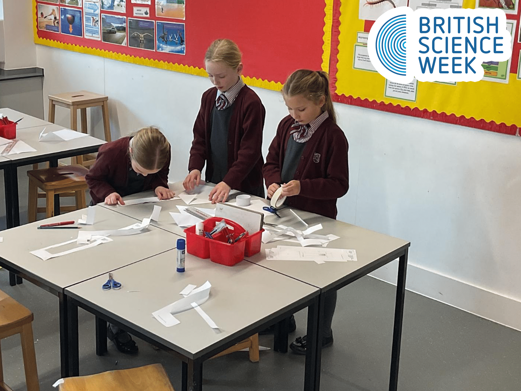 Three young girls in school uniforms work on science projects with paper and glue at a classroom table. The British Science Week logo is visible in the top right corner.