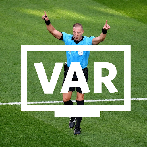 A football referee on the pitch signals with both hands raised, standing behind a bold white VAR graphic, indicating a Video Assistant Referee review during a match.