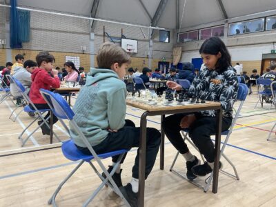 Children are seated at tables in a gymnasium, playing chess in pairs. The focus is on two players facing each other, concentrating on the game. Other children play in the background.