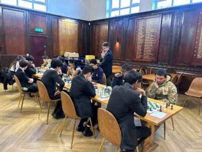 A group of students, mostly in school uniforms, play chess at wooden tables in a large hall. An adult stands observing. The room has wood-panelled walls and large plaques with names in the background.