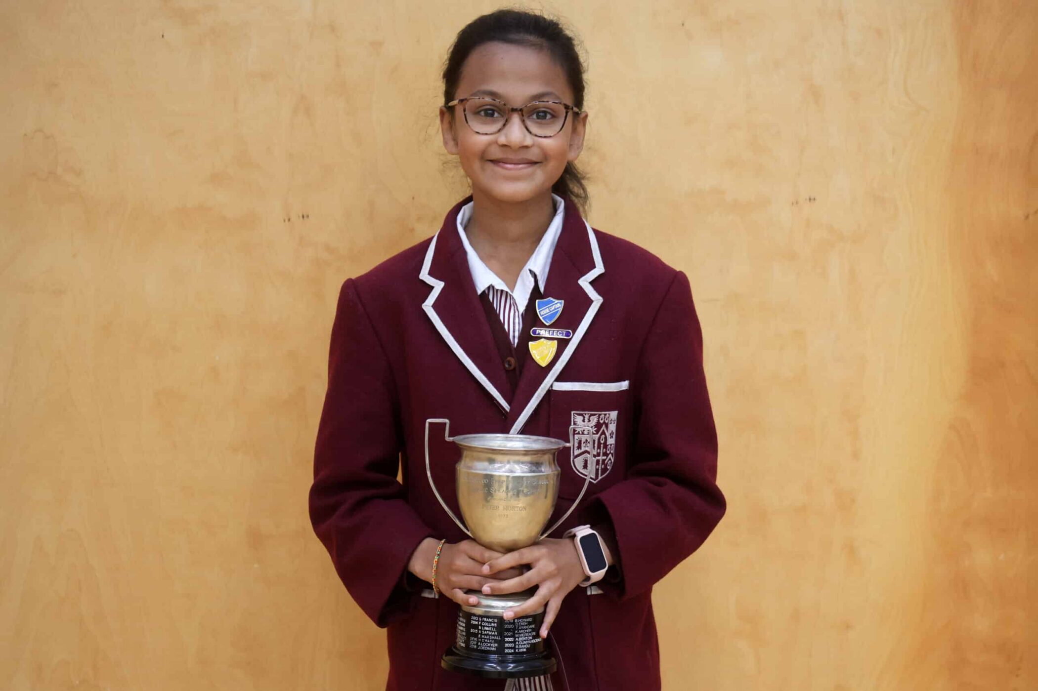 A young student in a maroon school blazer and glasses smiles whilst holding a silver trophy. She stands against a plain wooden background, displaying badges on her blazer.