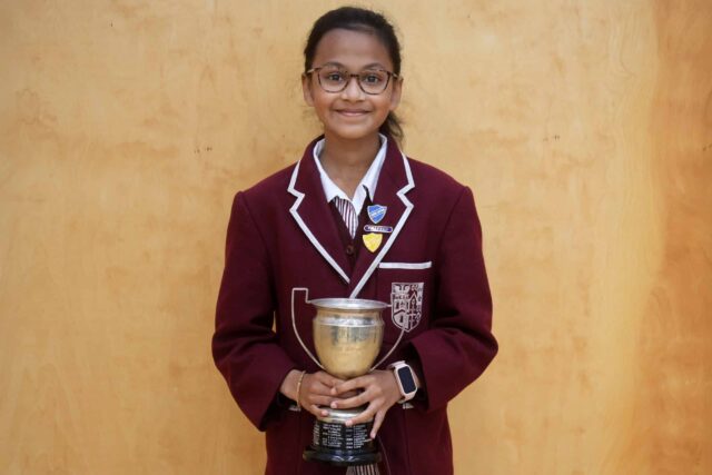 A young student in a maroon school blazer and glasses smiles whilst holding a silver trophy. She stands against a plain wooden background, displaying badges on her blazer.