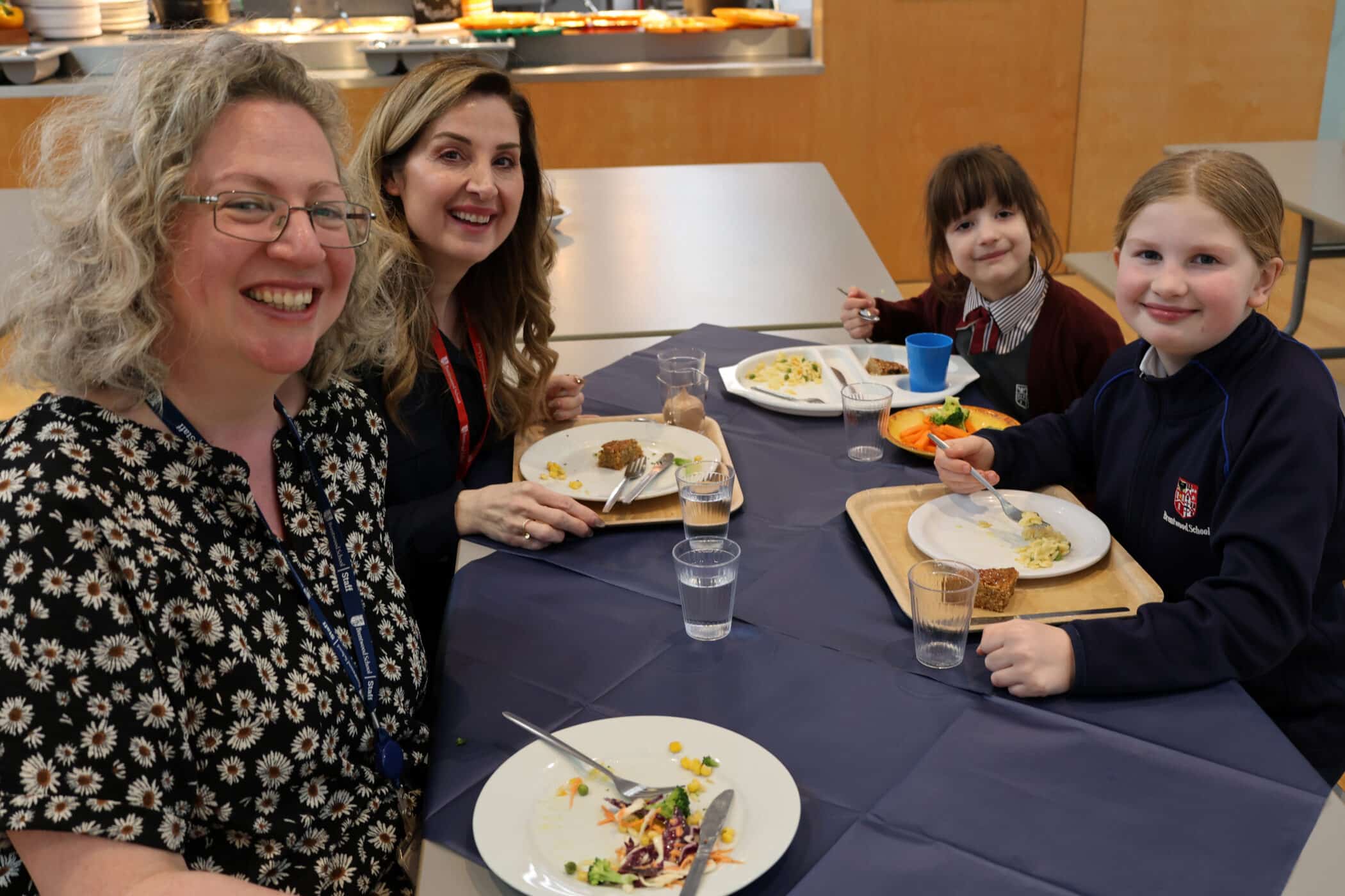 Four people—two adults and two children—are sitting at a table in a canteen, smiling at the camera while eating a meal. Plates of food, cups of water, and trays are on the table.