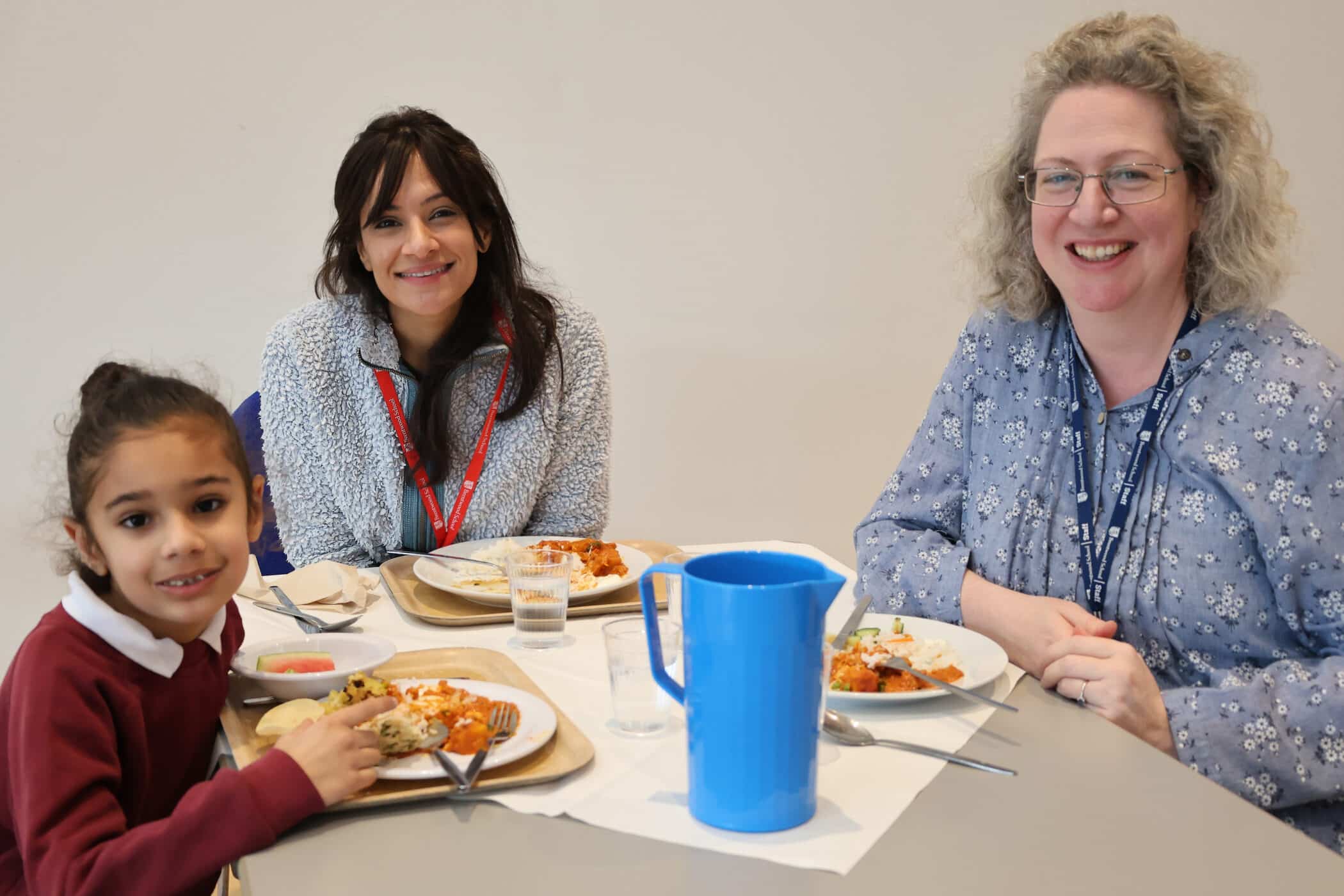 Three people, two adults and a child, sit at a table sharing a meal. The child smiles at the camera, whilst the adults also smile. Plates of food, cups, and a blue jug are on the table.